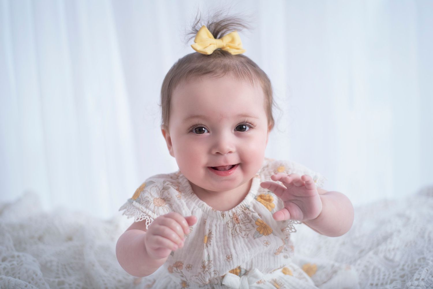 A happy baby in a white lace dress reaches up while sitting on soft white fabric.