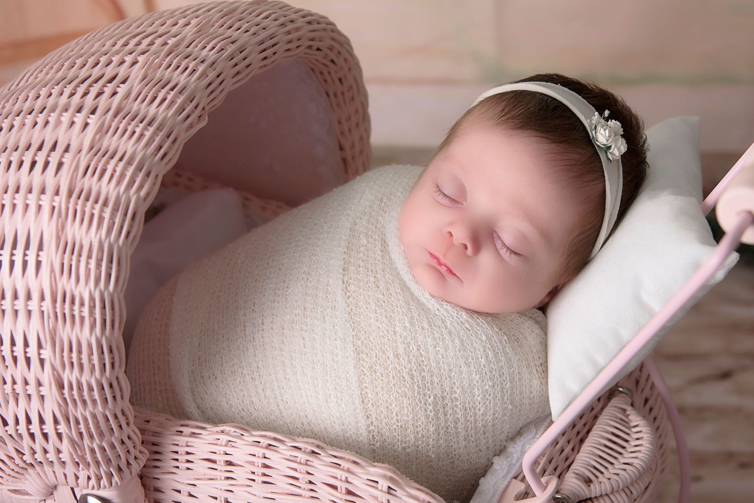 Newborn baby girl sleeping peacefully in a pink wicker bassinet, wrapped in cream knit, wearing a floral headband.