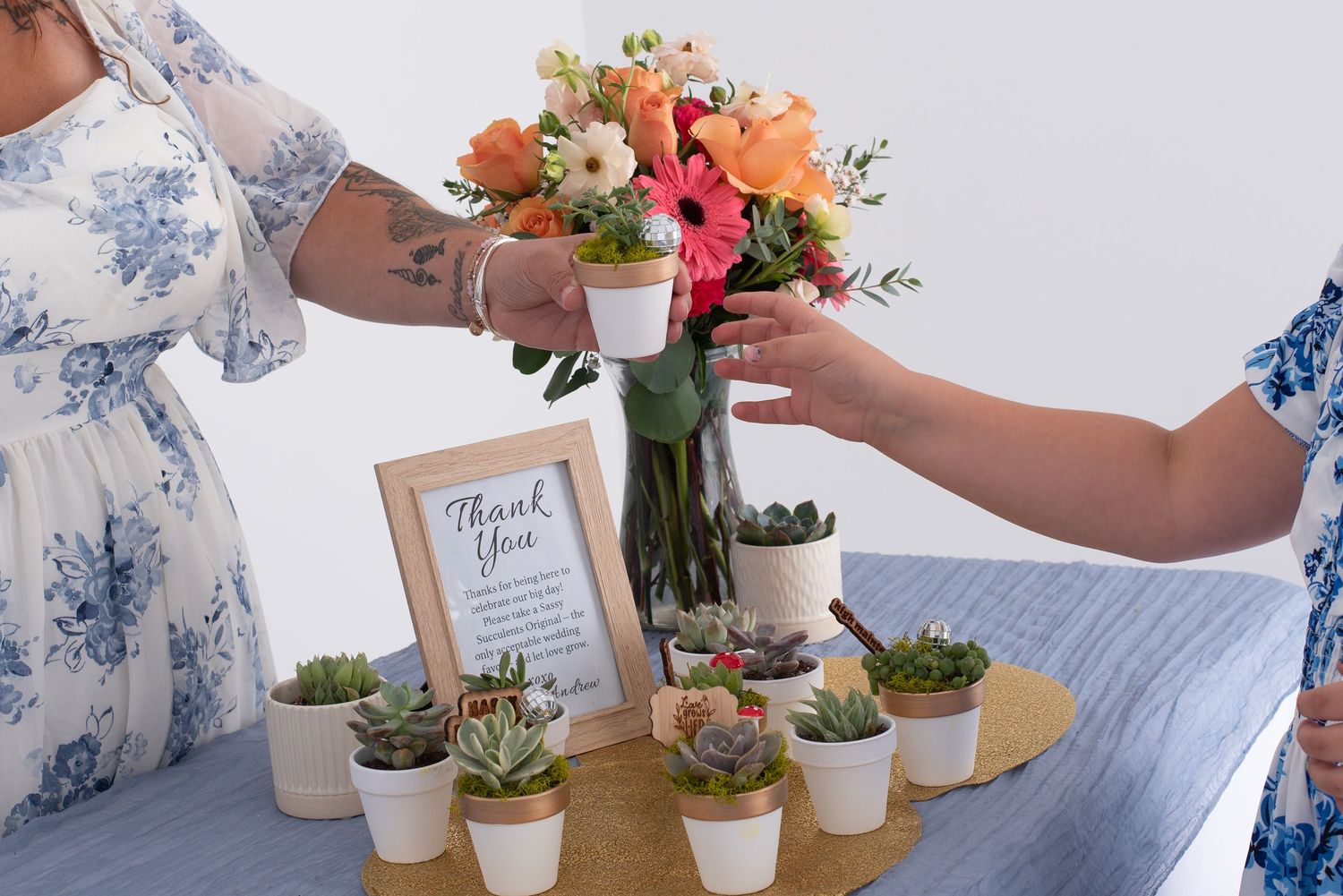 Hands arranging small potted succulent plants and floral centerpieces on a white table surface.
