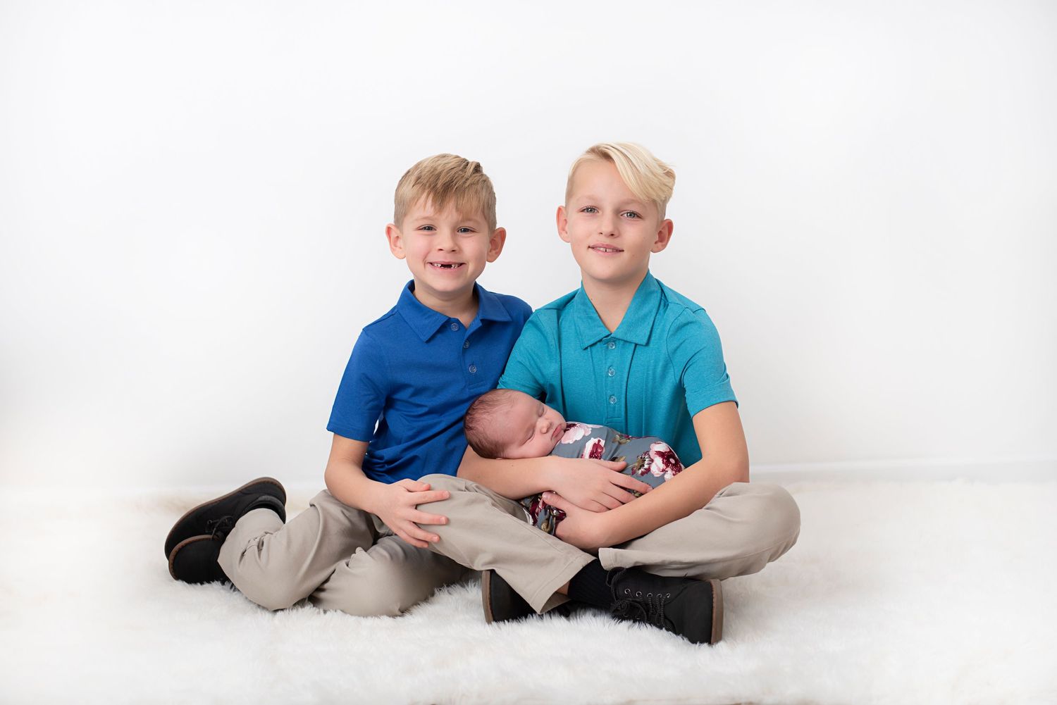 Two young siblings in blue polo shirts sitting cross-legged on a white backdrop with a newborn baby. Rhode Island Photographer