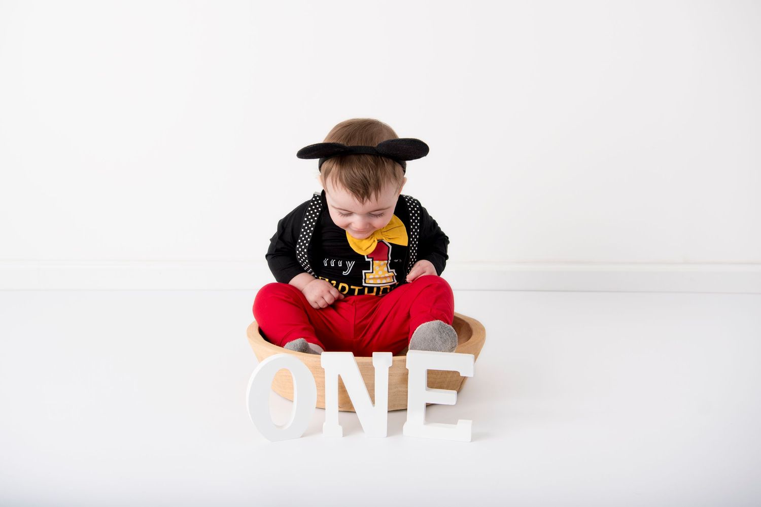Baby wearing black and red outfit with black hat sits next to white wooden 'ONE' letters on white backdrop.