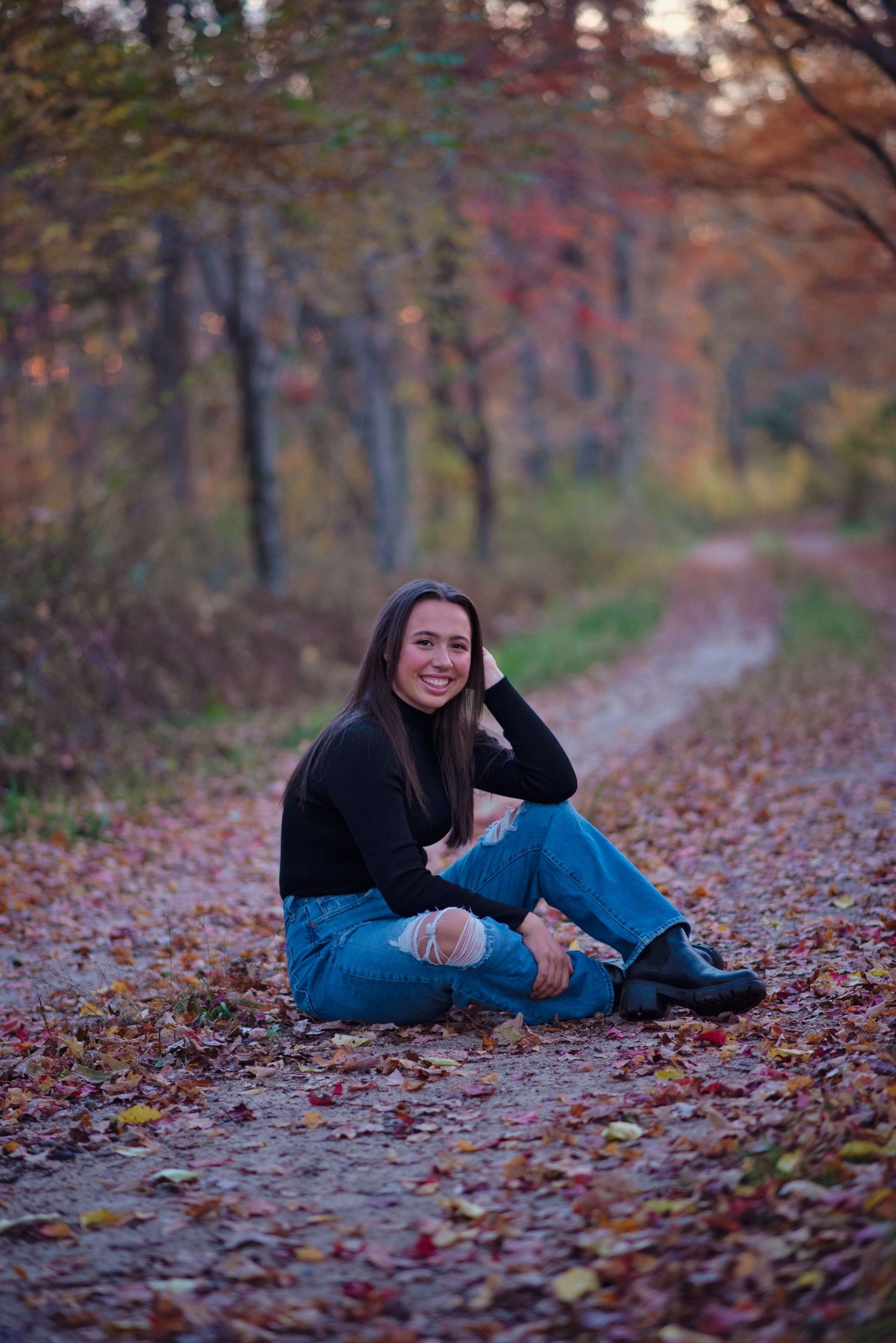 Someone in distressed jeans sits casually on a leaf-covered forest path during autumn with colorful trees in the background.