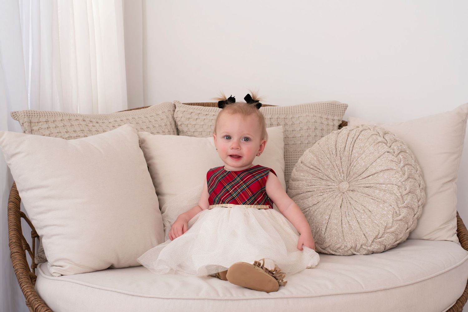 Small child in red plaid and white dress sitting on cream colored couch with decorative throw pillows.