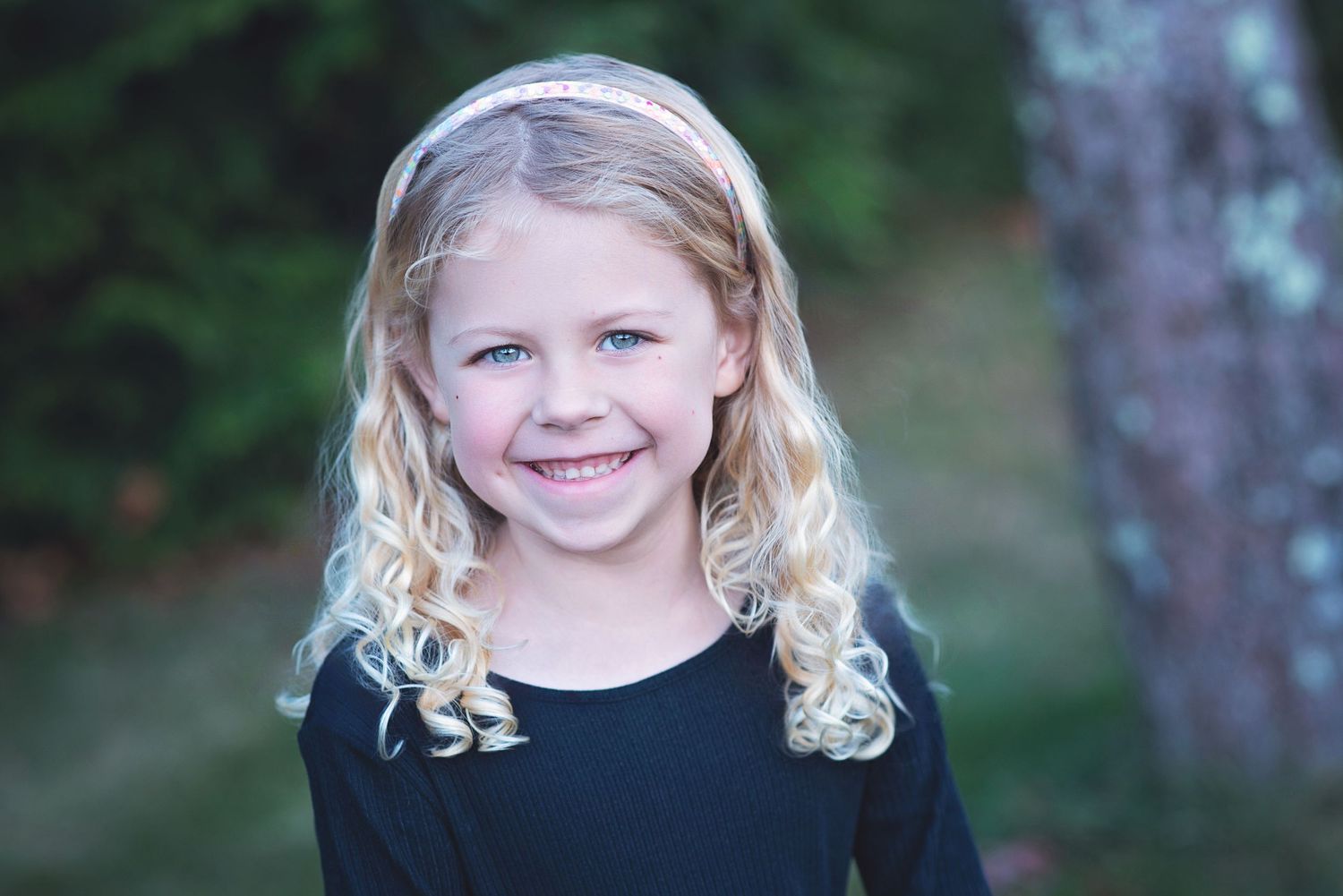 A child with curly blonde hair wearing a black shirt smiles warmly outdoors against a blurred green background. Exeter, RI Photographer