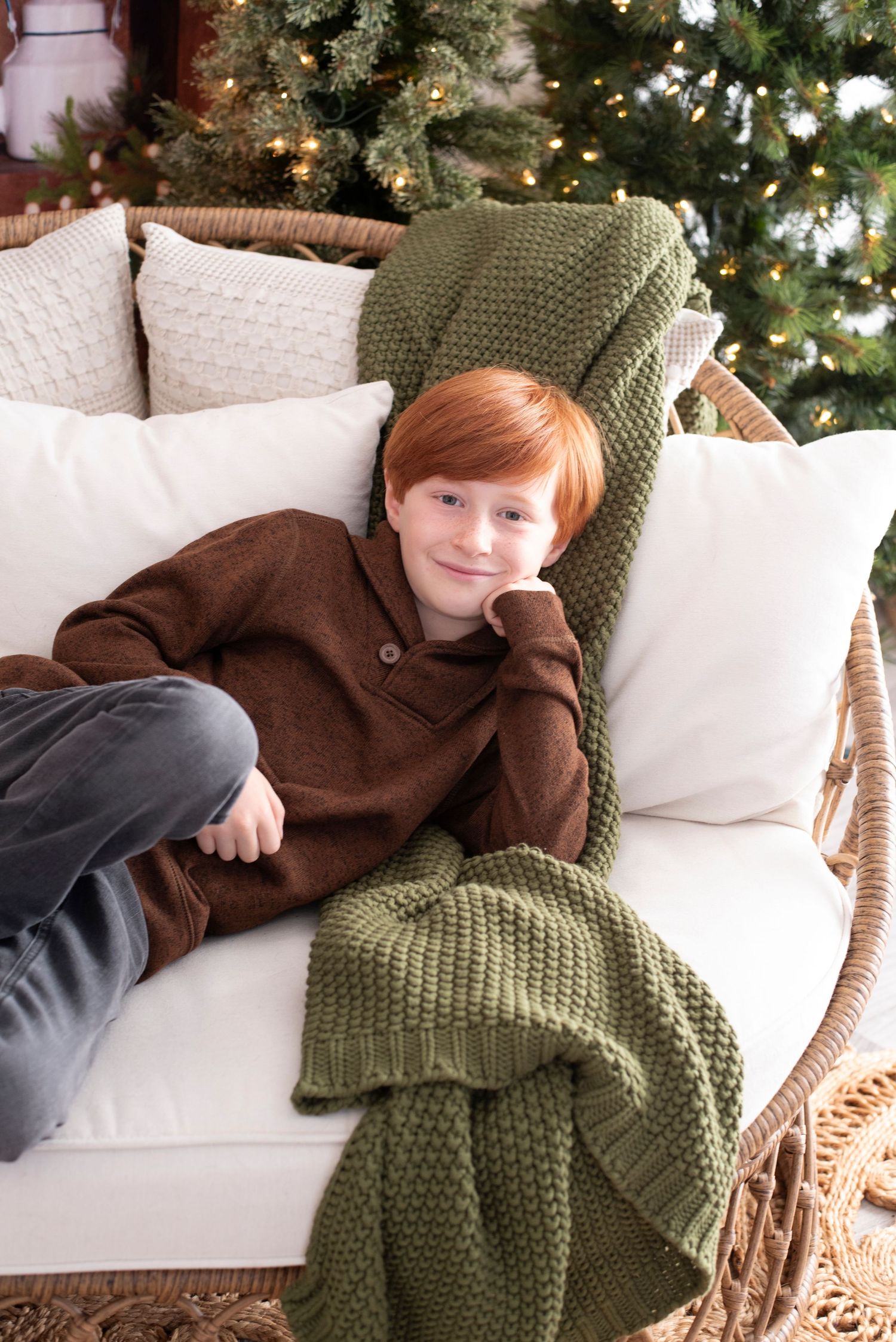 A person relaxes on a white couch with a dark brown sweater and olive green knit throw blanket near a Christmas tree.