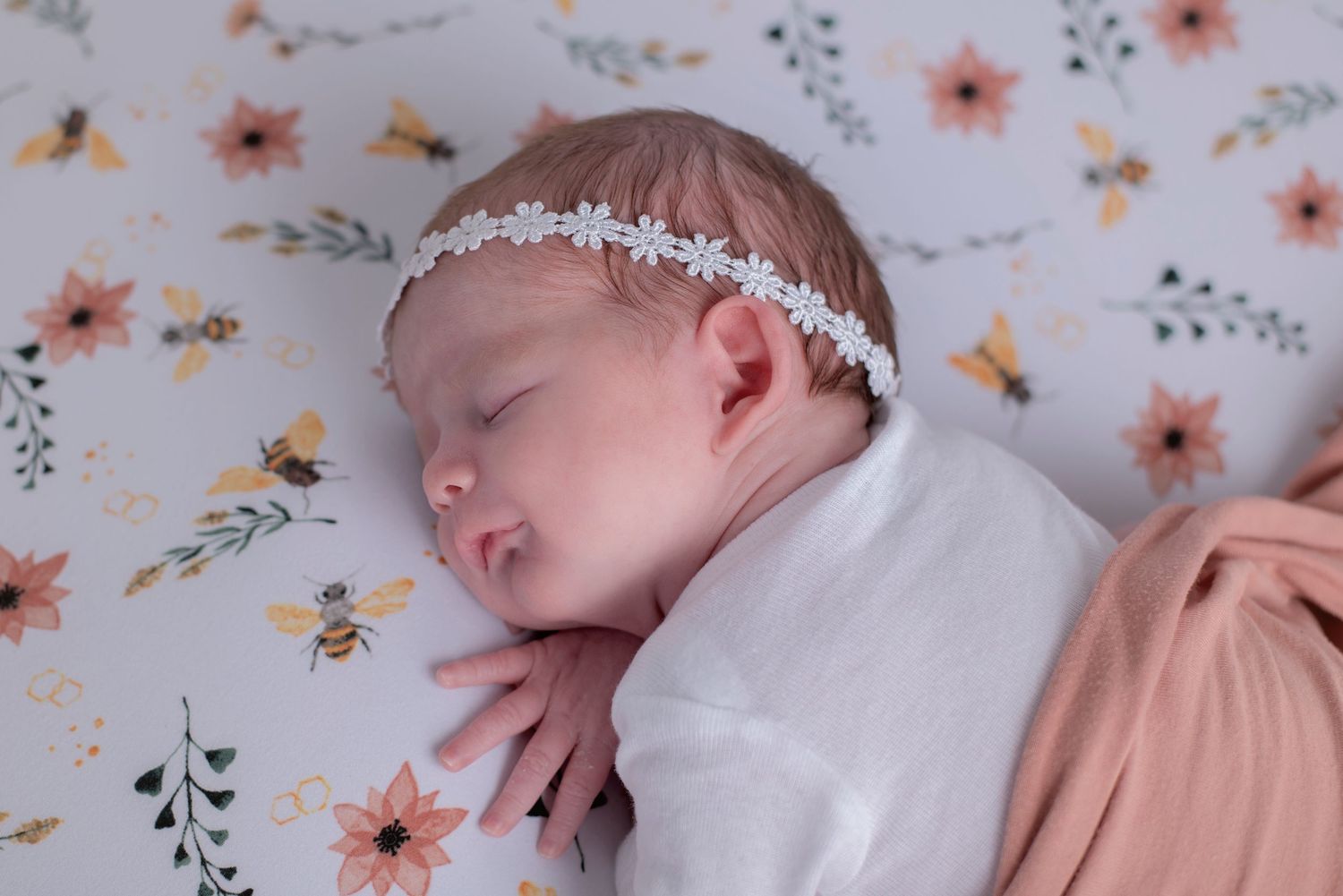 A sleeping newborn baby wearing a delicate white headband rests on a floral printed fabric.