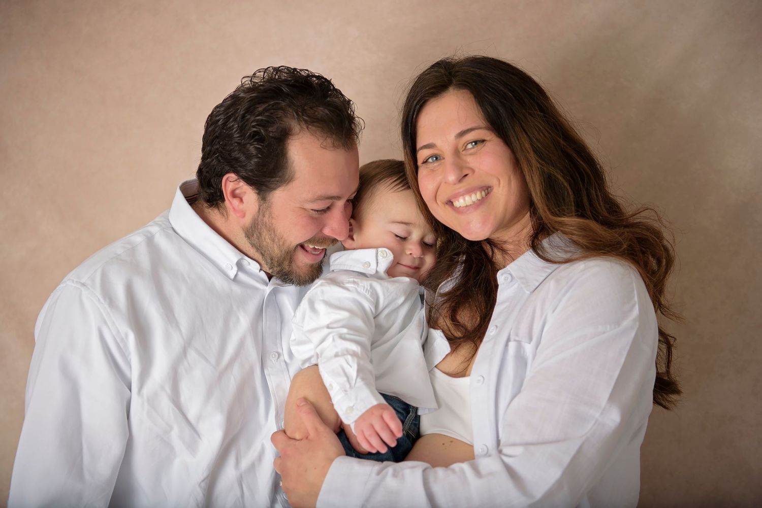 Happy family portrait with parents in white shirts smiling at their baby against a warm beige background.