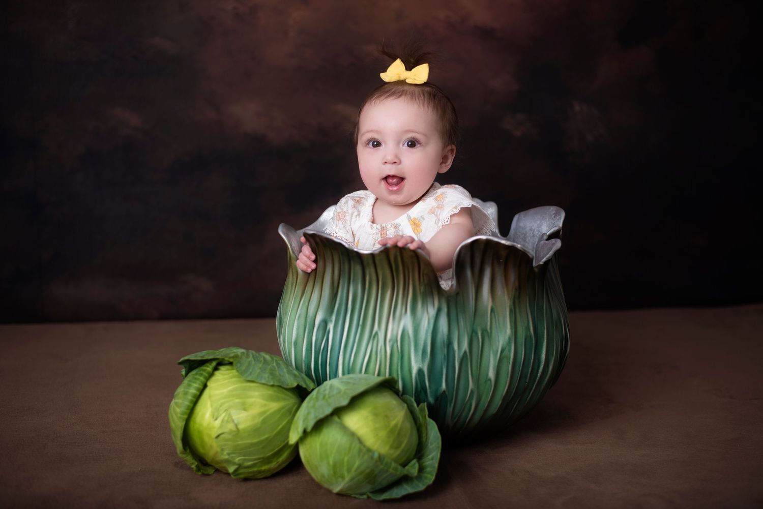 Baby wearing a yellow bow sits nestled inside green cabbage leaves during a whimsical studio portrait session.