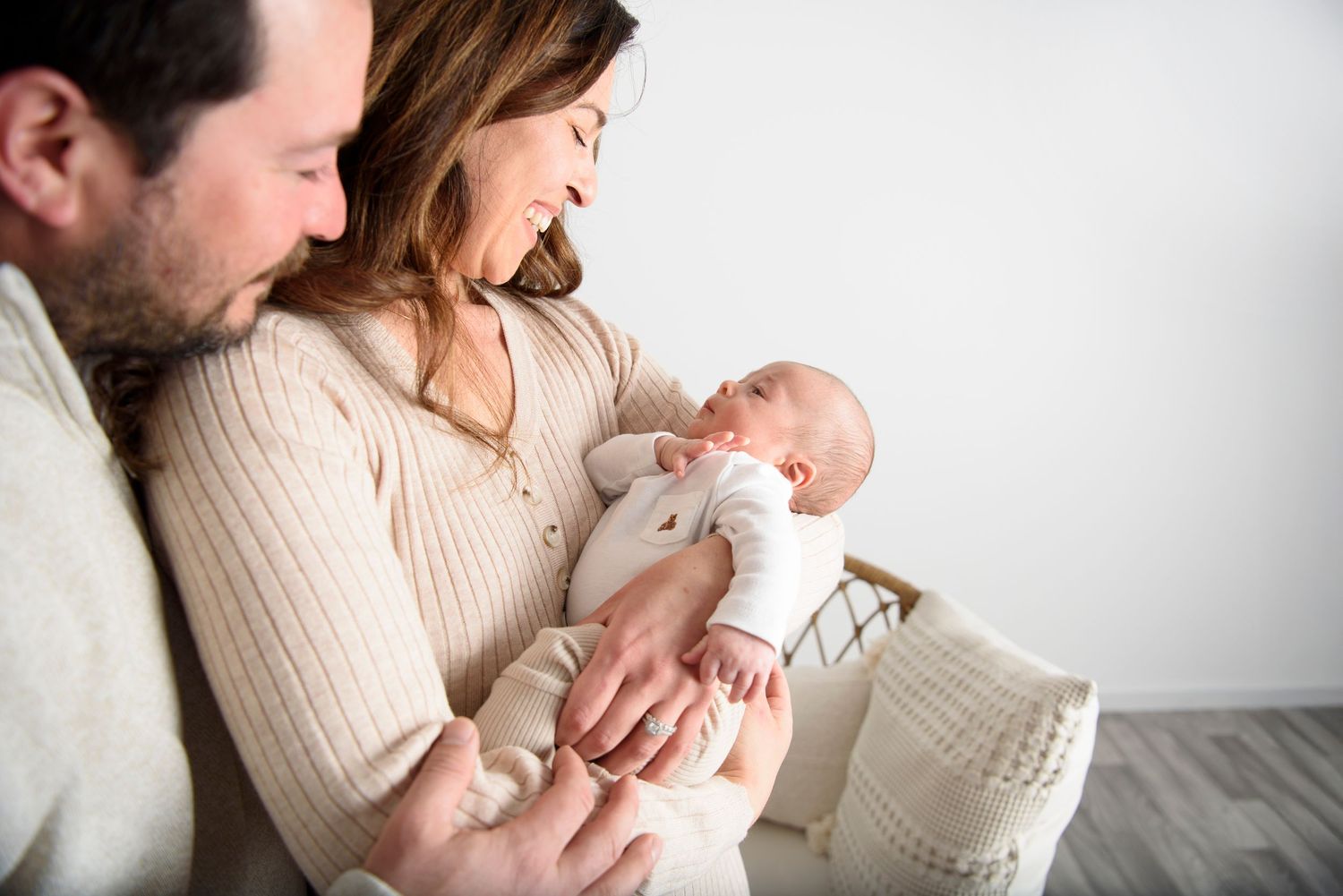 Parents in cream sweaters lovingly hold their newborn baby against a white backdrop.