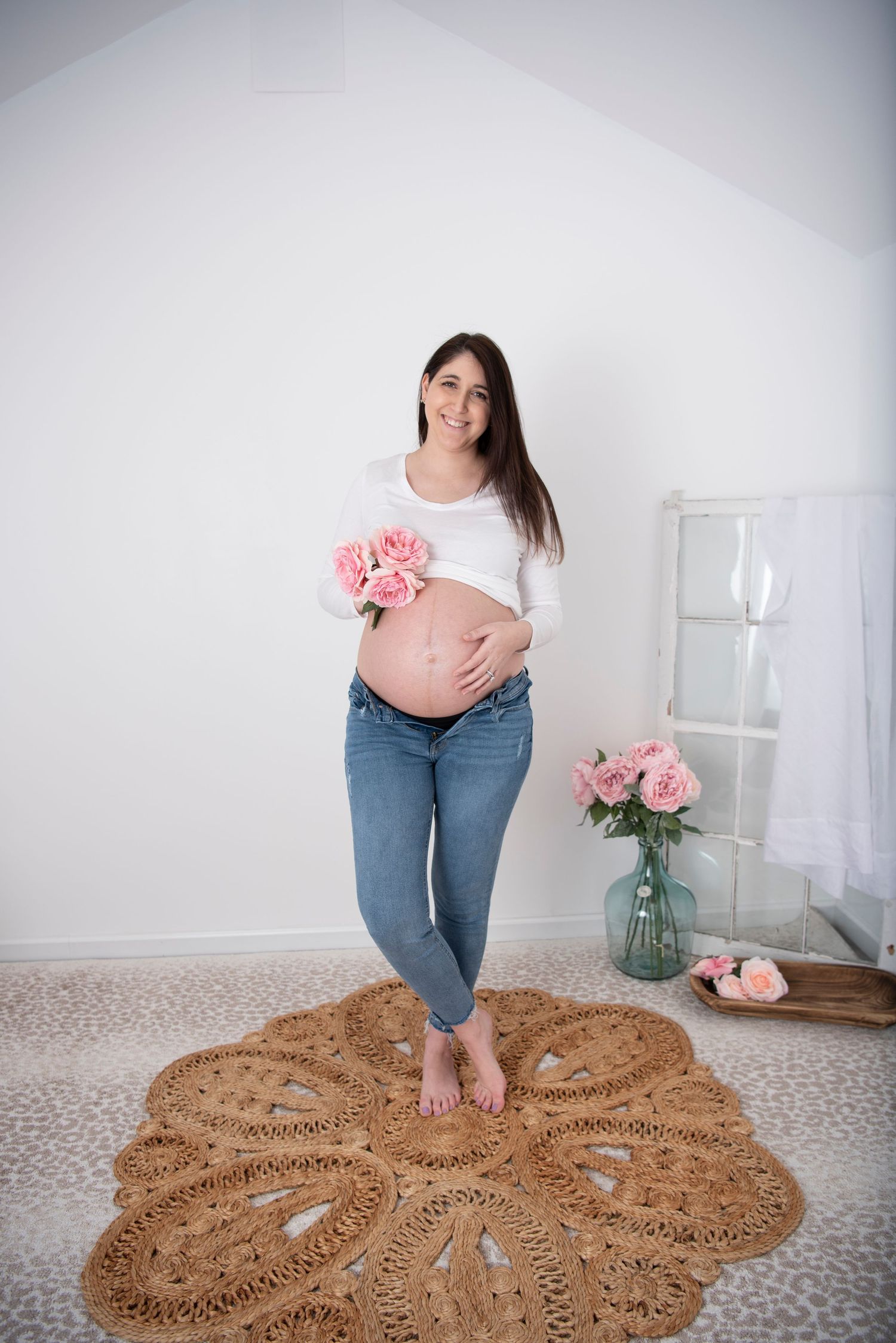 Expectant mother in jeans and white top stands barefoot on round brown rug holding pink flowers in minimalist setting.