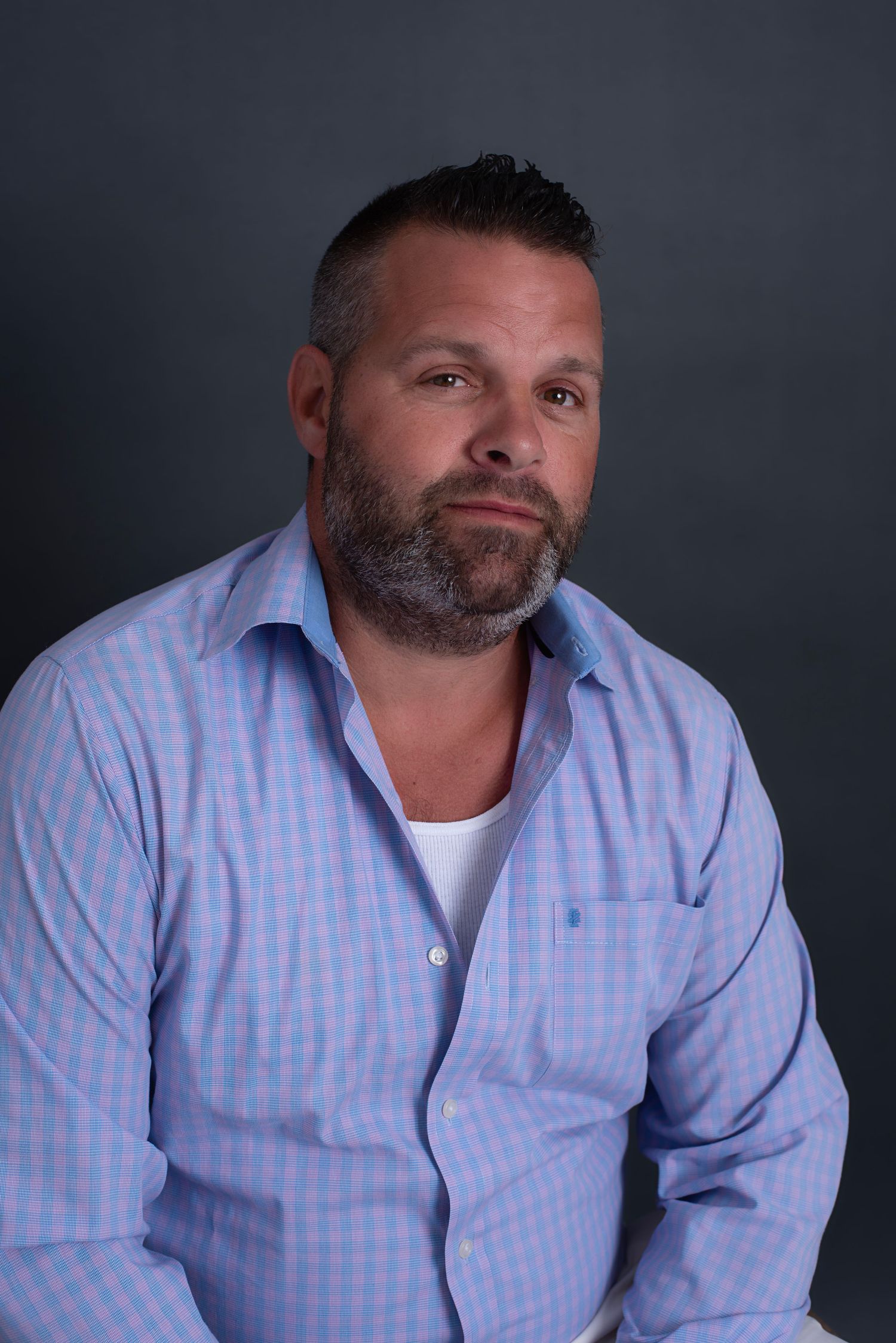 Professional studio portrait against dark background showing person in light blue checkered button-down shirt and white undershirt.