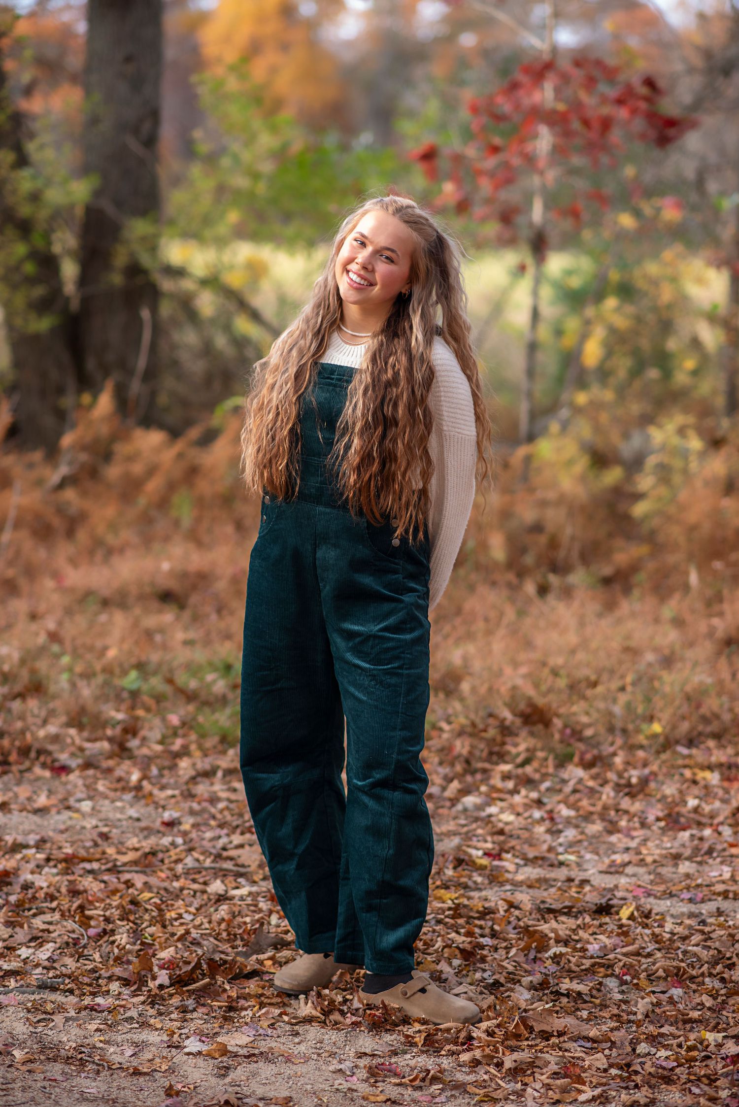 Long-haired person in dark green overalls poses joyfully among autumn foliage on a forest trail. Rhode Island Photographer