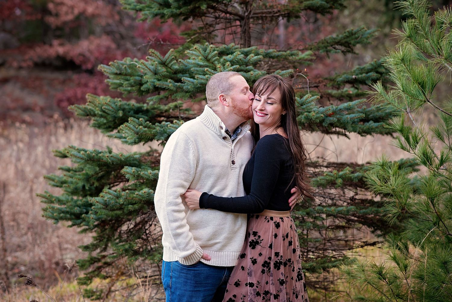 A romantic couple shares intimate moments among pink cherry blossoms and evergreen trees in a spring garden setting.