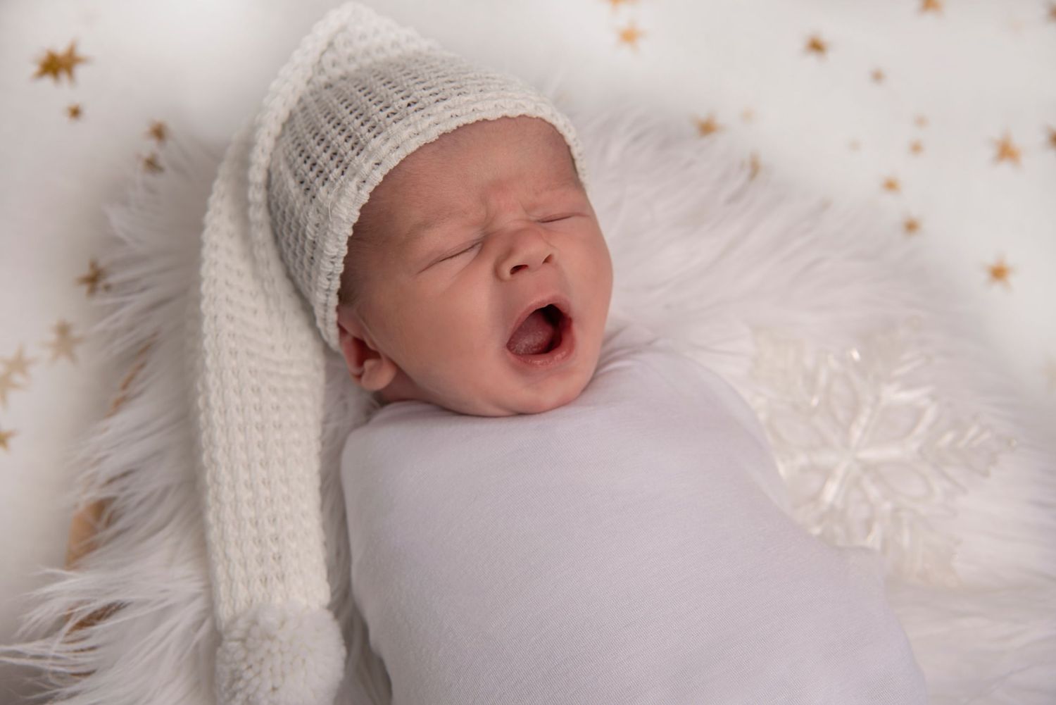 Newborn baby yawning while wearing white knit hat and swaddled in white blanket on fur background with gold stars.