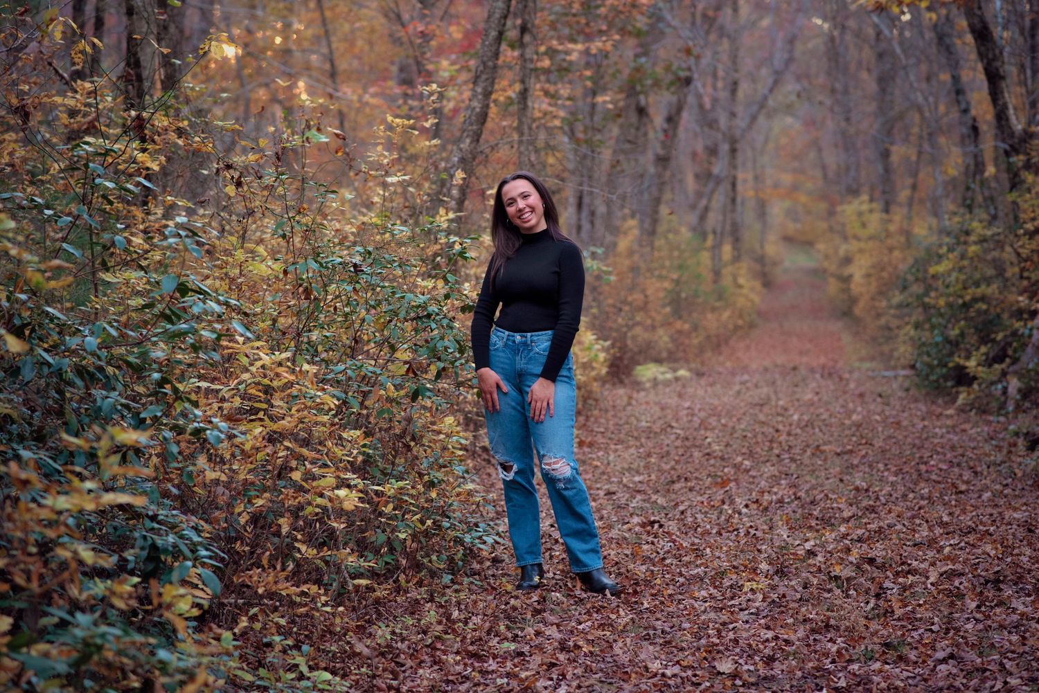 Someone poses on a leaf-covered forest path during autumn with golden foliage surrounding the trail.
