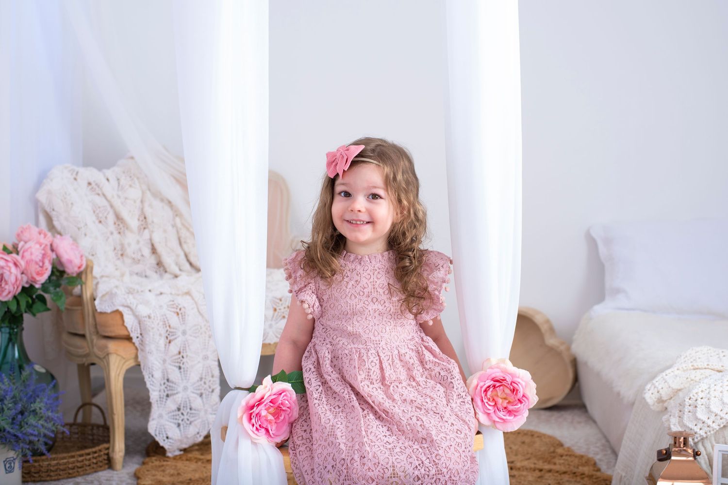 A child in a pink lace dress poses in a white room decorated with pink roses and white lace.