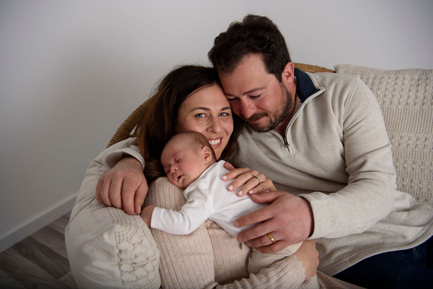 A peaceful family moment as parents gently embrace their sleeping newborn while sitting on a cozy chair at home.