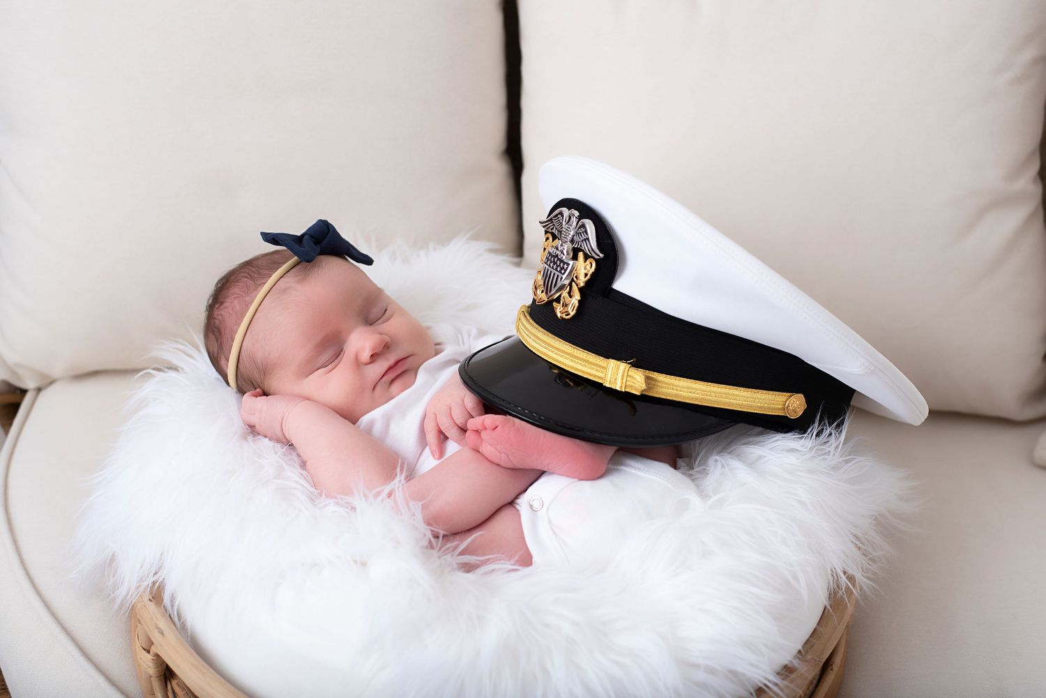 Newborn baby wrapped in white blankets sleeps on fluffy white fur while wearing a navy sailor hat with gold brim.