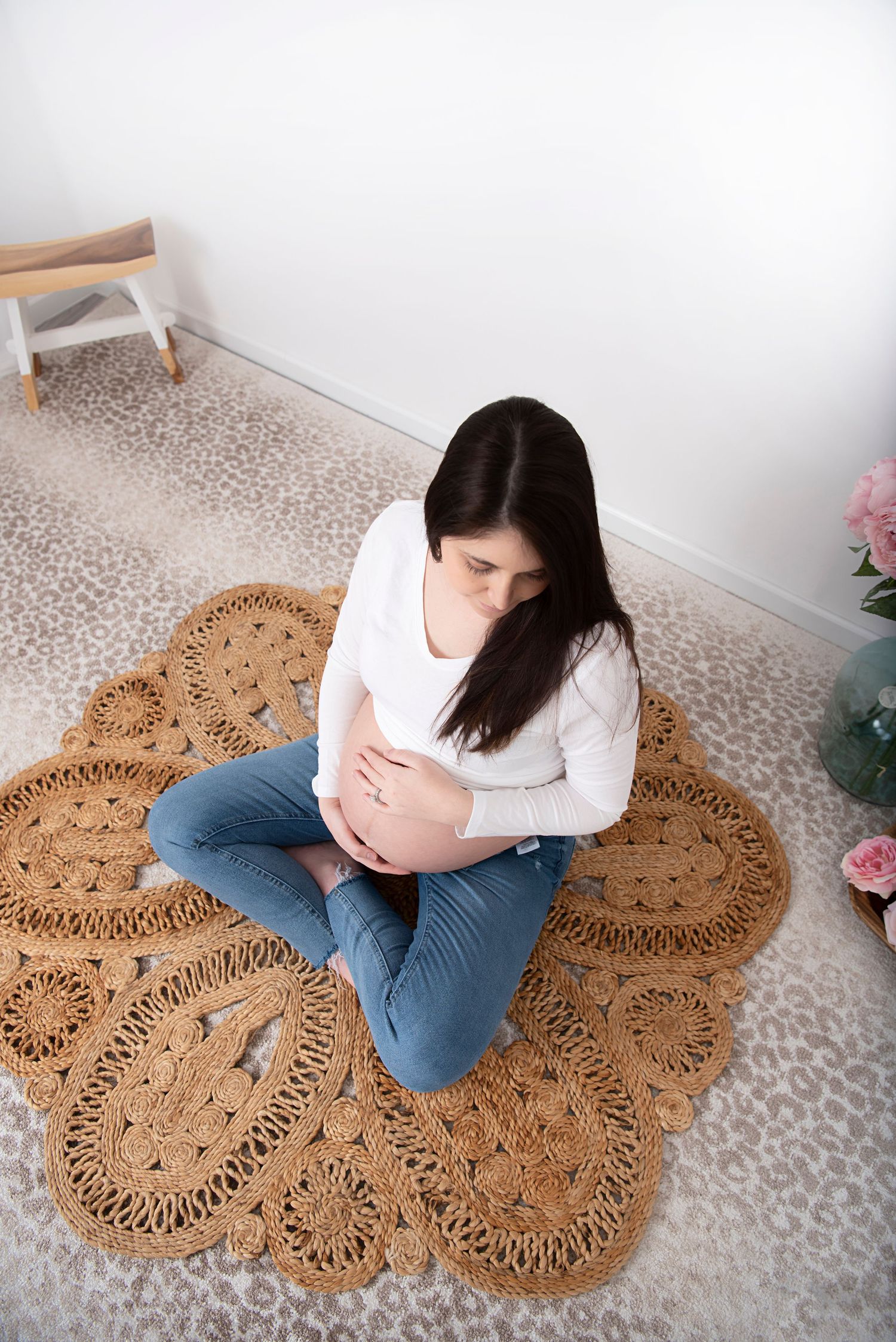 Person sits cross-legged on decorative jute floor rug while cradling pregnant belly in white sweater and jeans.