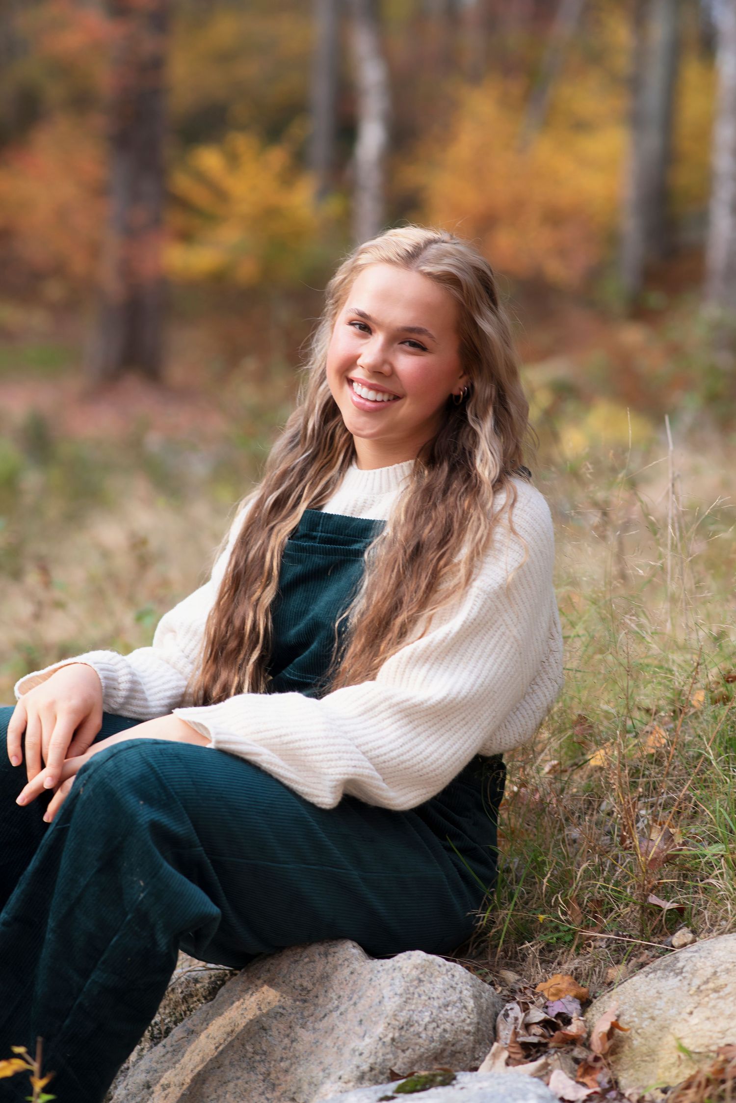 Person in white knit sweater and green corduroy overalls sits on rocks amidst autumn foliage.