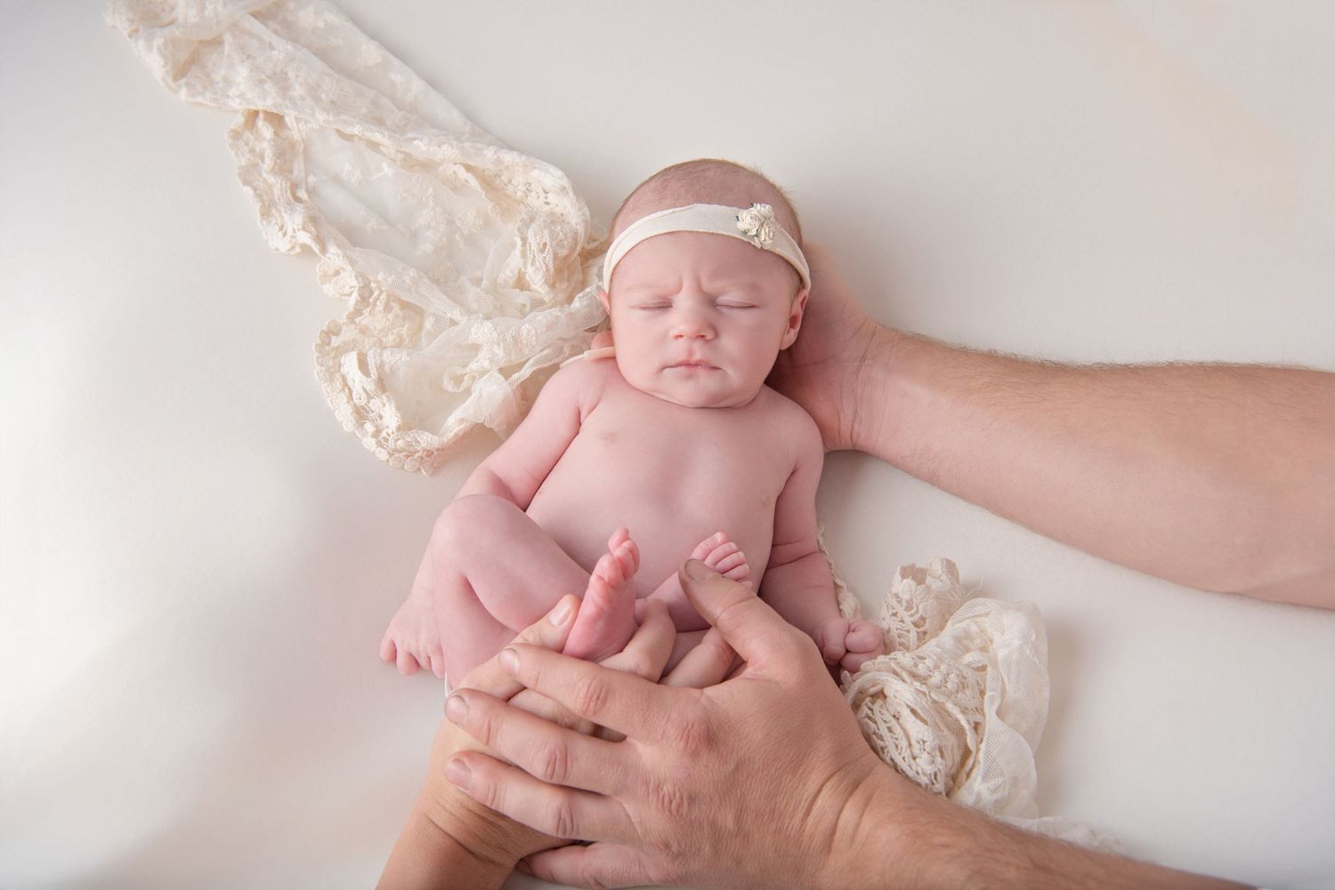 A newborn baby lies peacefully on white lace fabric while being gently cradled by adult hands.