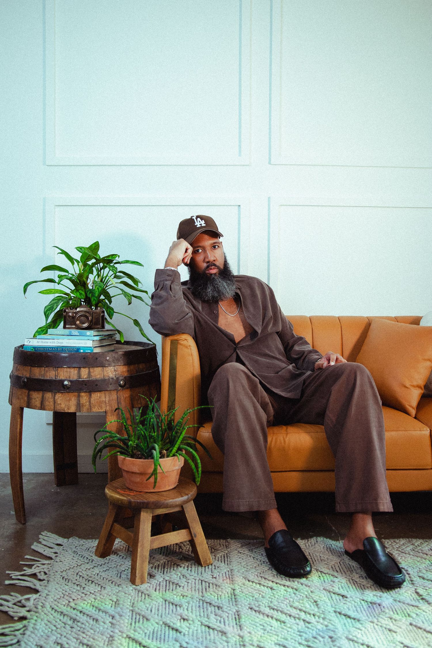 Bearded man in brown suit and LA cap sits on tan leather sofa beside barrel table with plants.