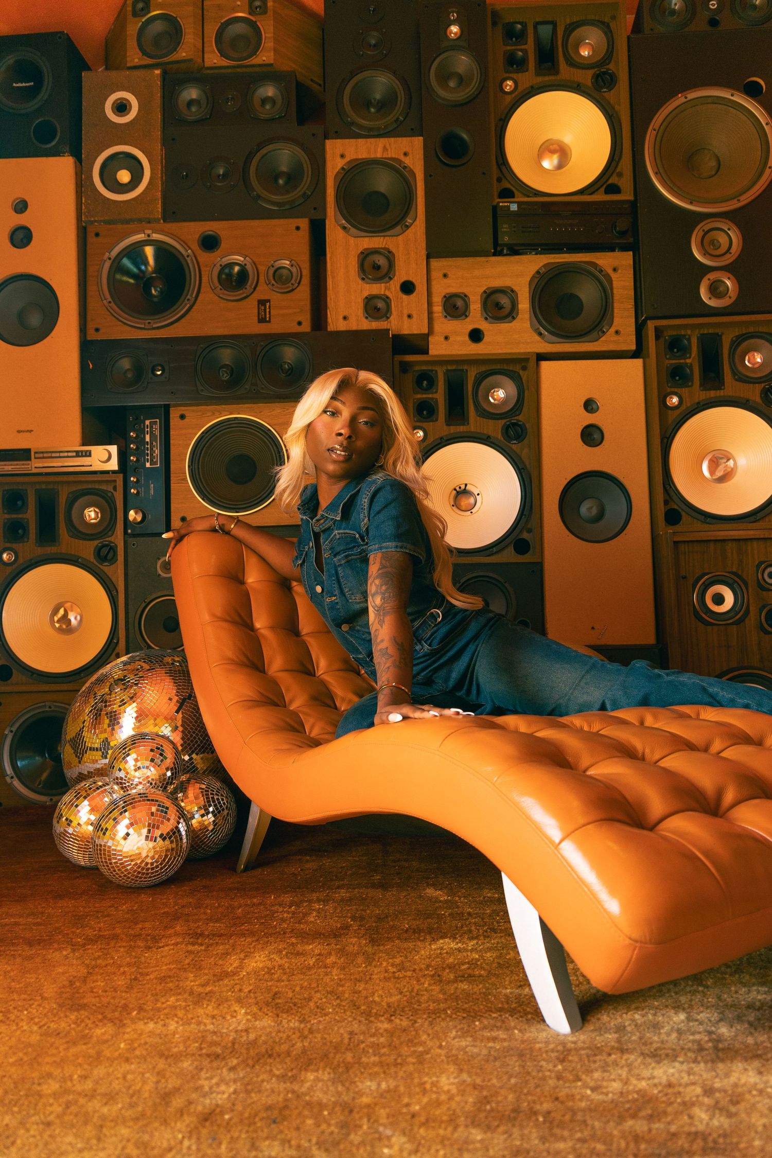 Tattooed woman in blue denim reclines on orange chaise lounge with disco balls against wall of vintage speakers.