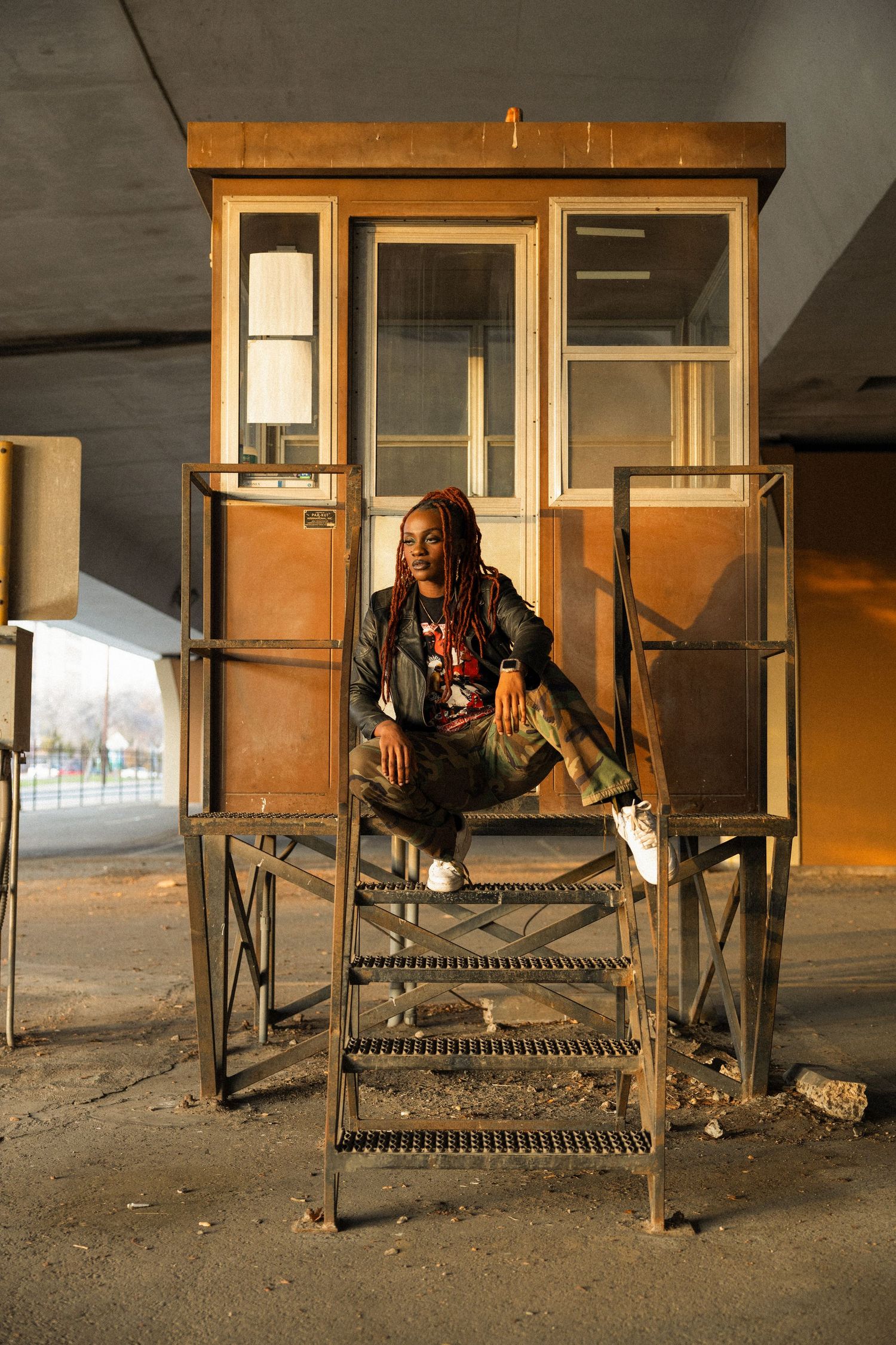 Person with dreadlocks sitting on metal stairs in front of vintage booth in urban setting.