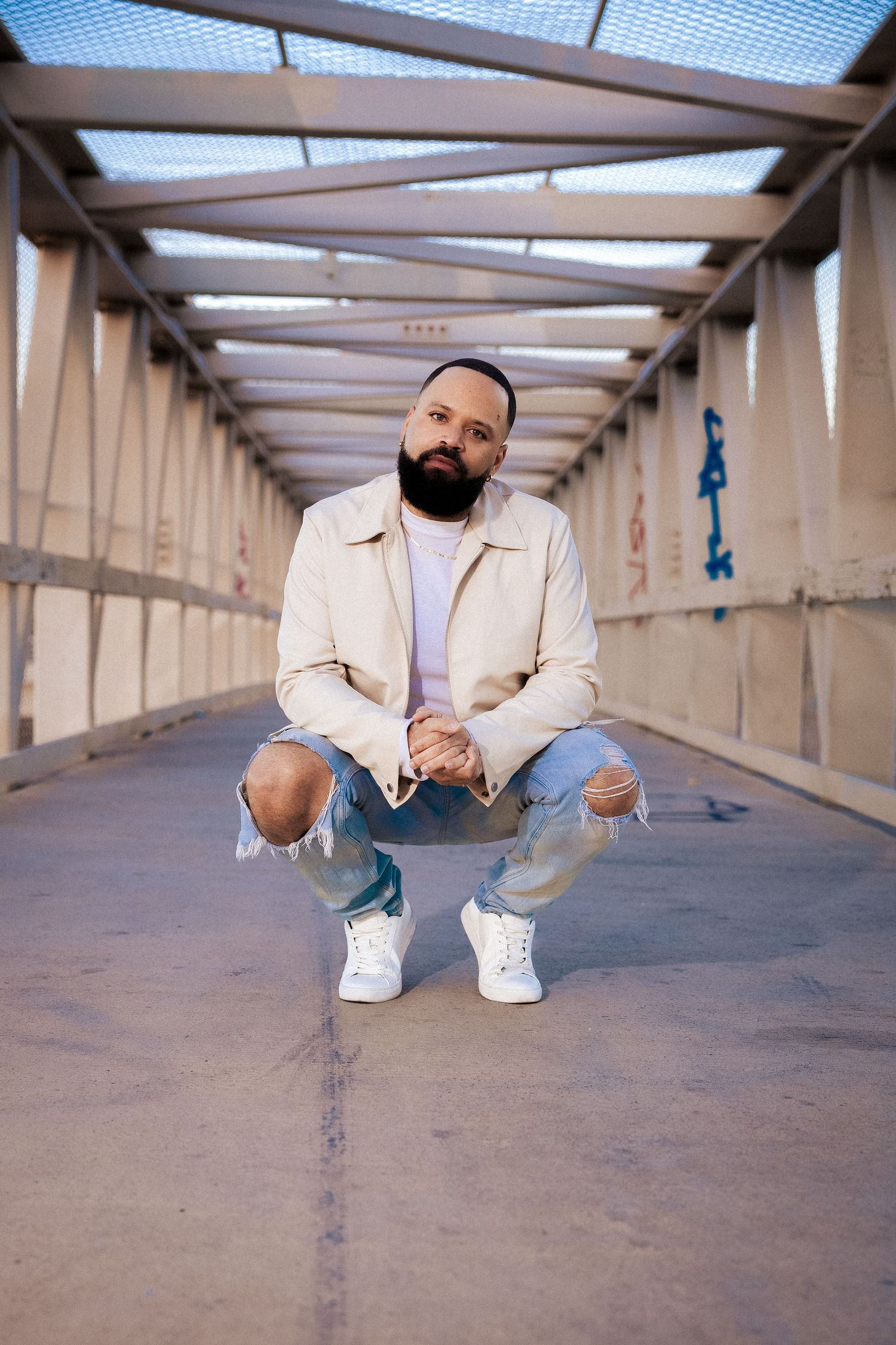 Bearded man squatting on urban bridge walkway wearing ripped jeans and white sneakers.