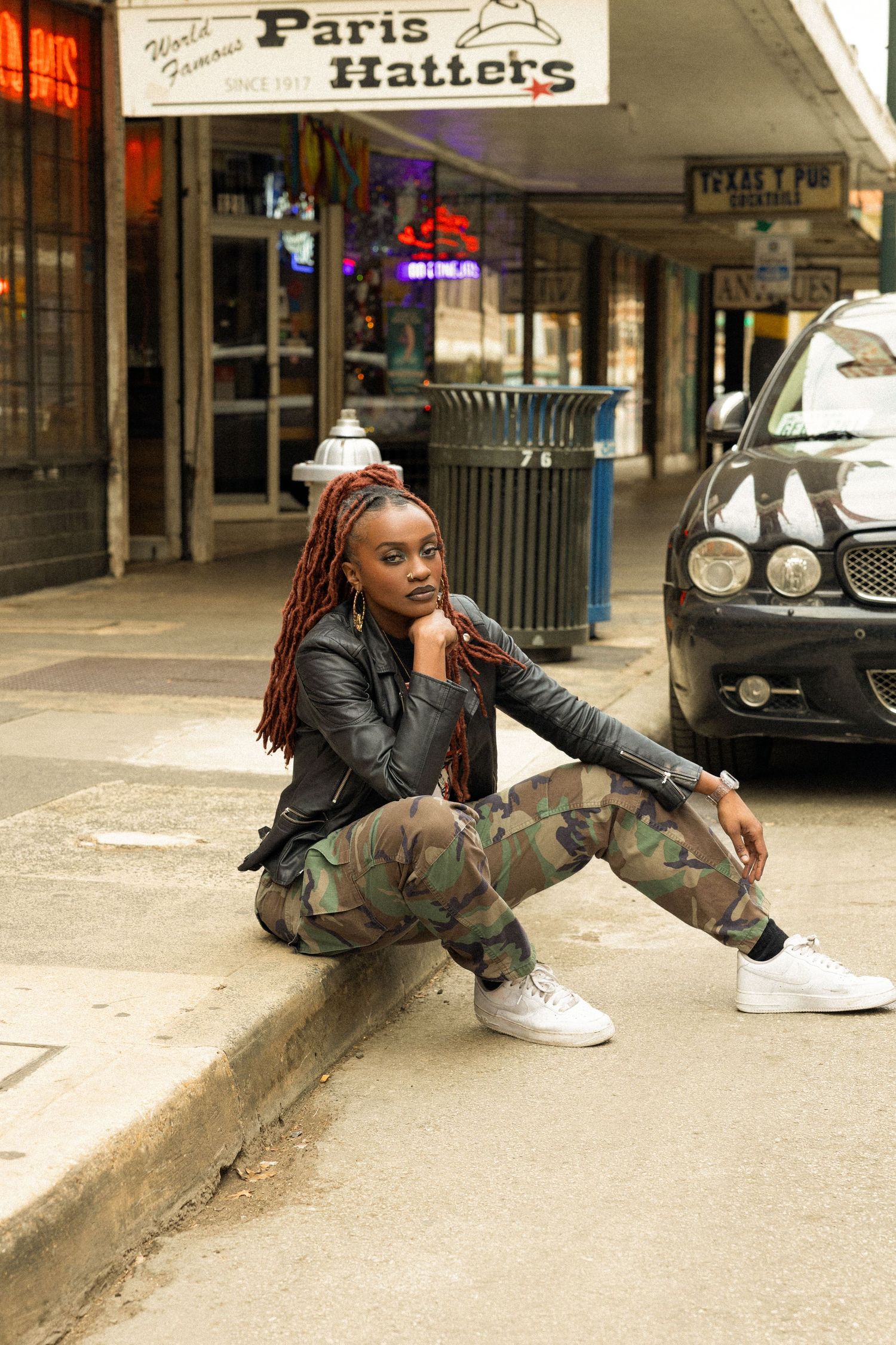 Woman with red braids in camo pants sits on urban curb near shops.