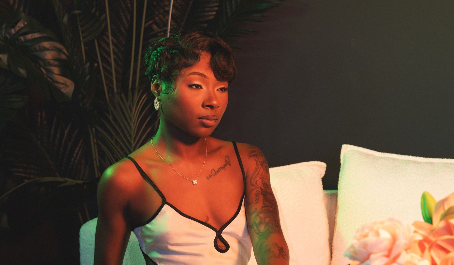 Woman with short hair sits pensively under neon green light near plants.