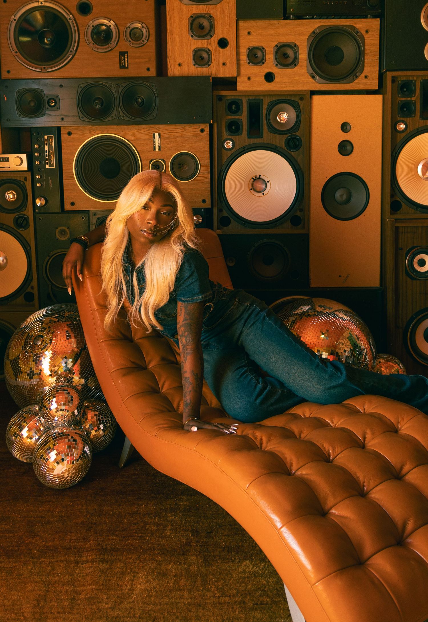 Woman with long blonde hair lounges on curved orange tufted chaise with disco balls and vintage speaker wall behind.