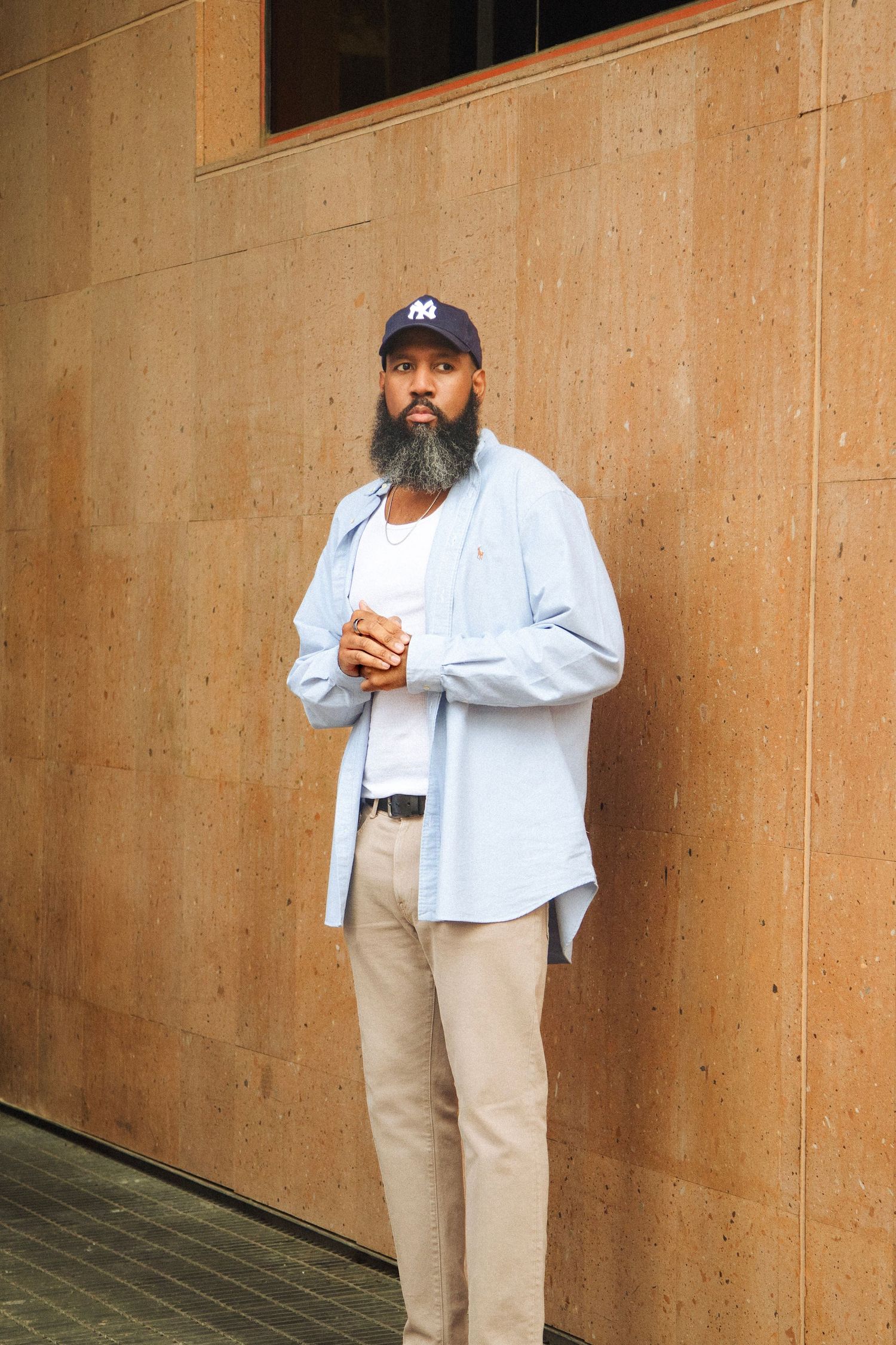 Bearded man in blue oxford shirt and khakis holds coffee cup while leaning against textured cork wall outdoors.