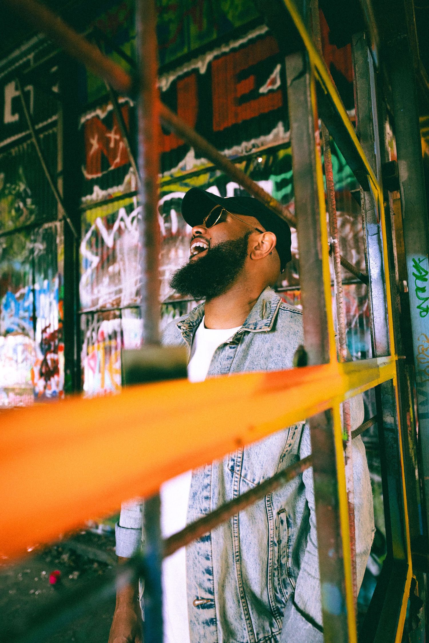 Bearded man in denim jacket and sunglasses laughing near colorful graffiti wall with orange scaffolding.