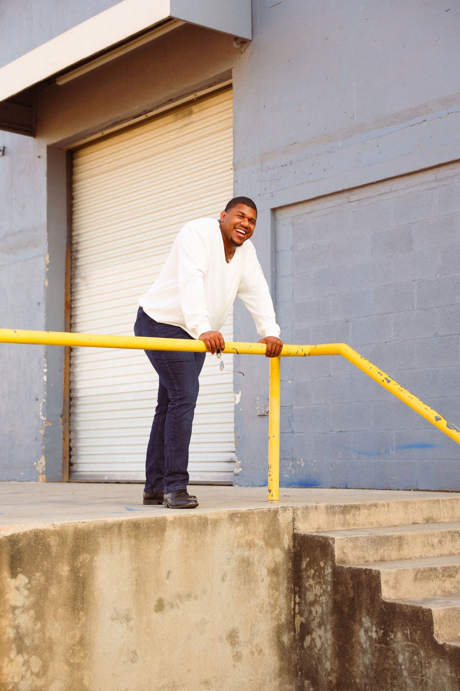Smiling man in white shirt leaning on yellow railing outside an industrial building with roll-up doors.