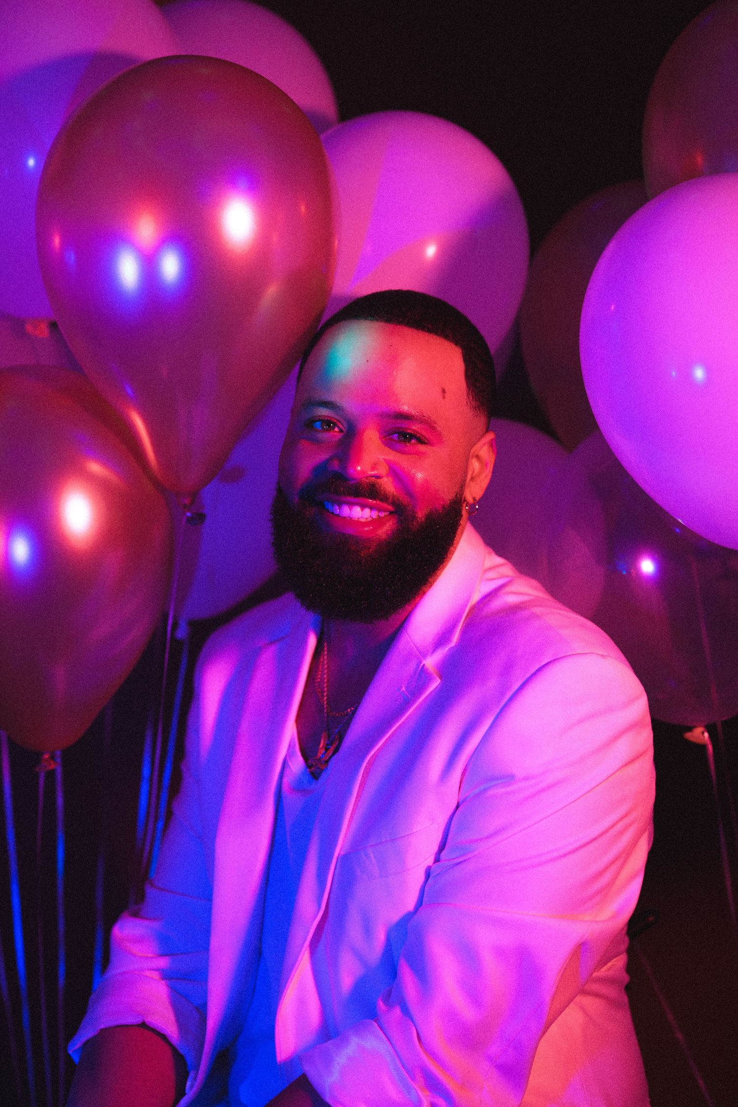 A smiling bearded man in a white blazer poses among metallic pink and purple balloons under neon light.