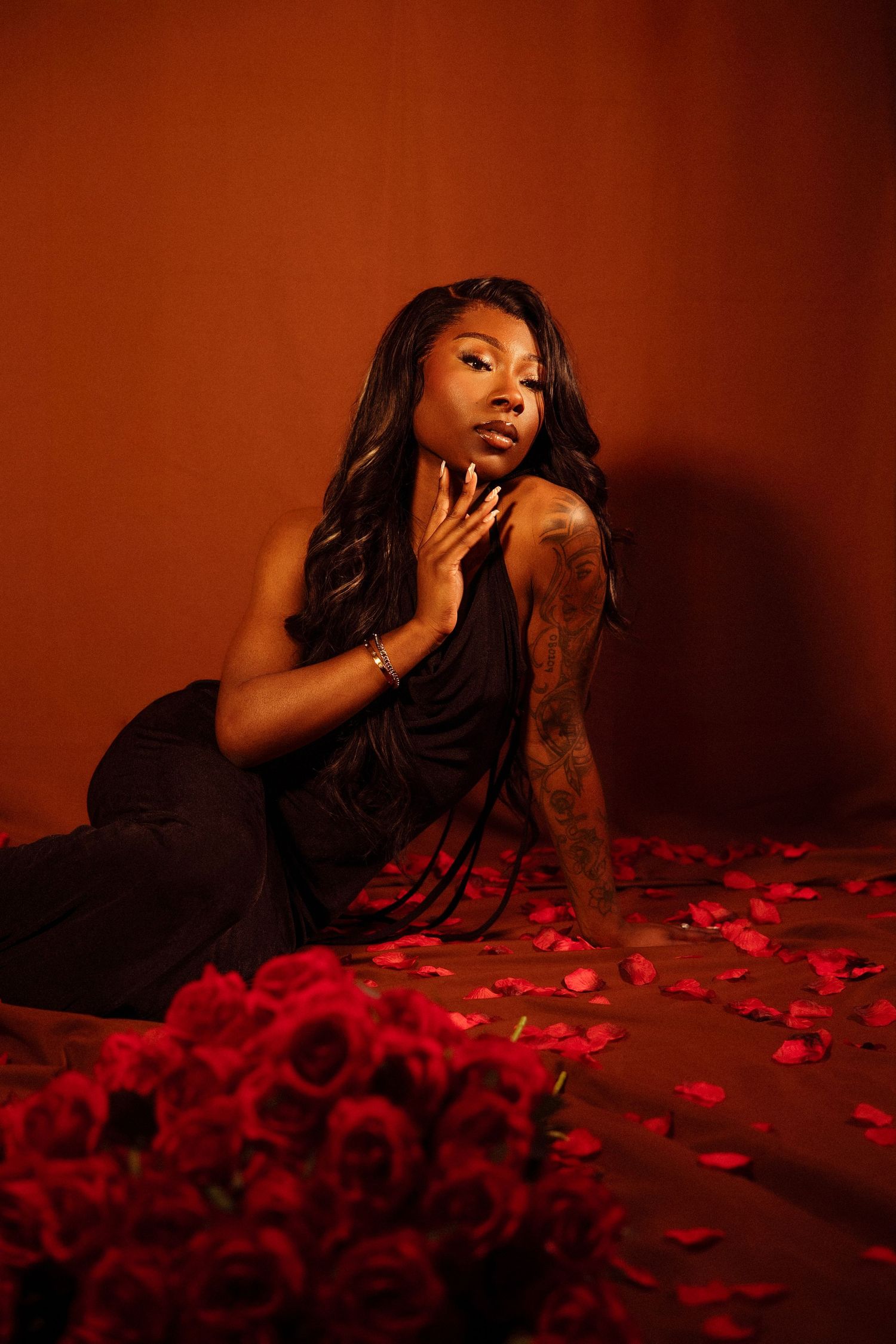 Tattooed woman in black halter dress seated among red rose petals against a rich red studio backdrop.