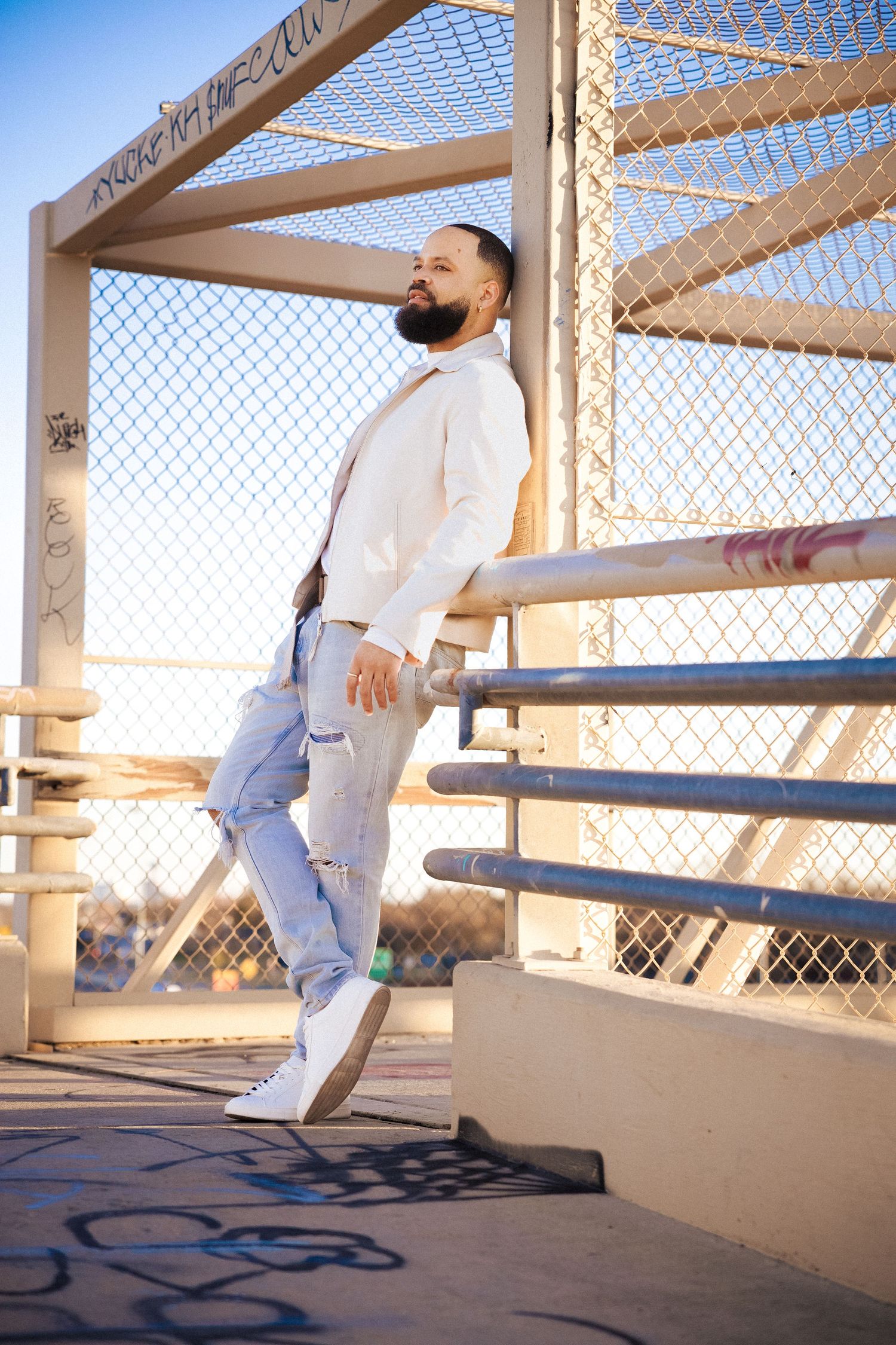 Stylish bearded man leaning on metal railing of urban bridge in white hoodie and ripped jeans.