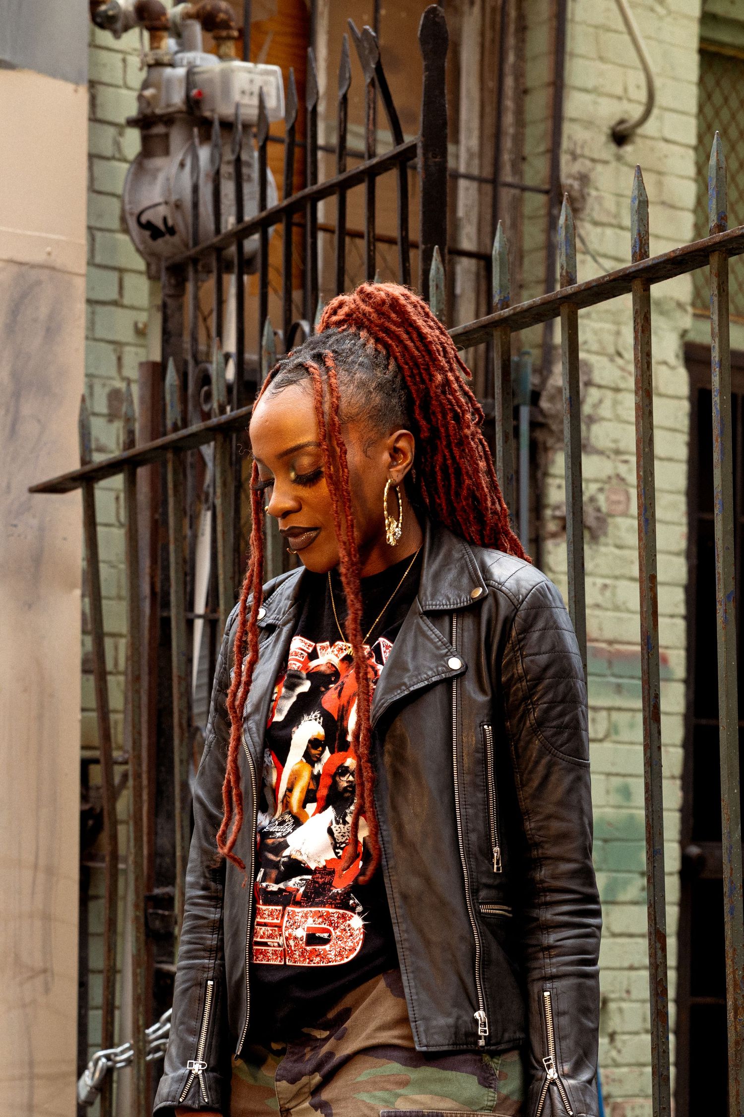 Black woman with long red dreadlocks wears leather jacket and graphic tee while standing near iron gate outdoors.