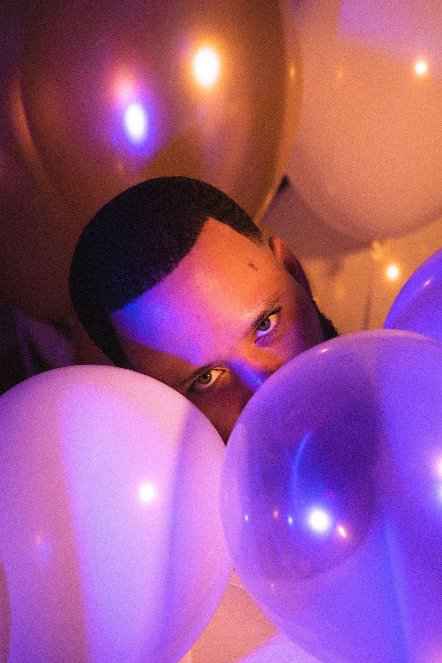 Person peeking through purple and gold balloons in dramatic purple lighting.