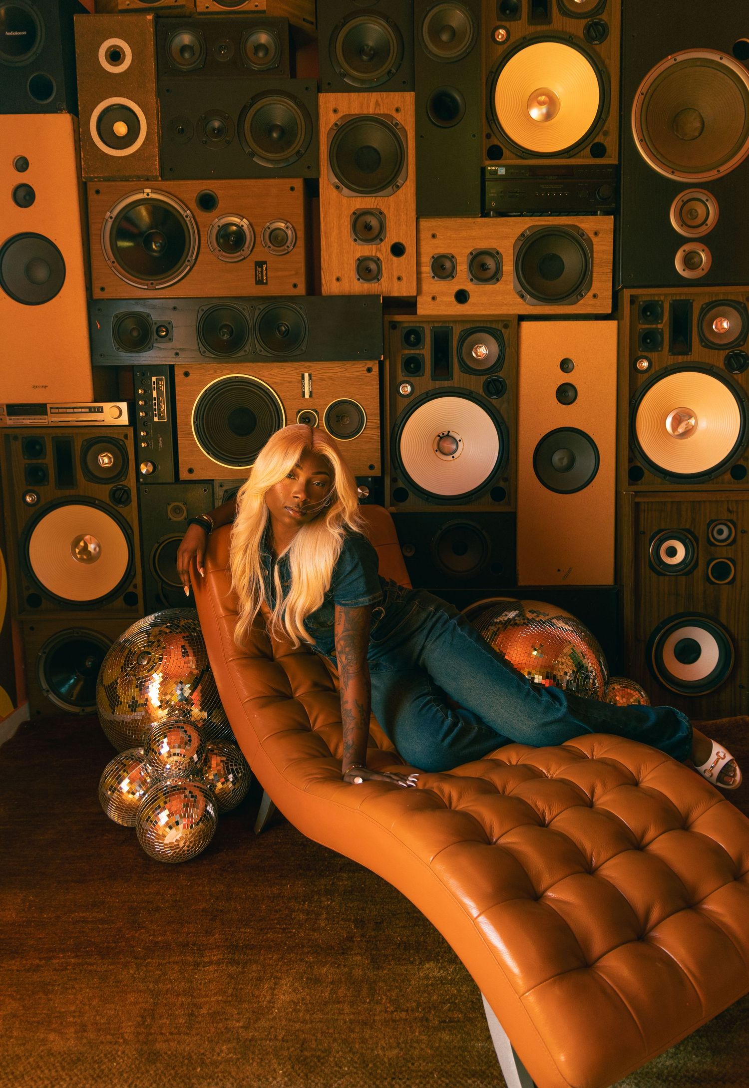 Blonde woman in jeans and boots reclines on orange tufted chaise with disco balls against retro speaker wall.