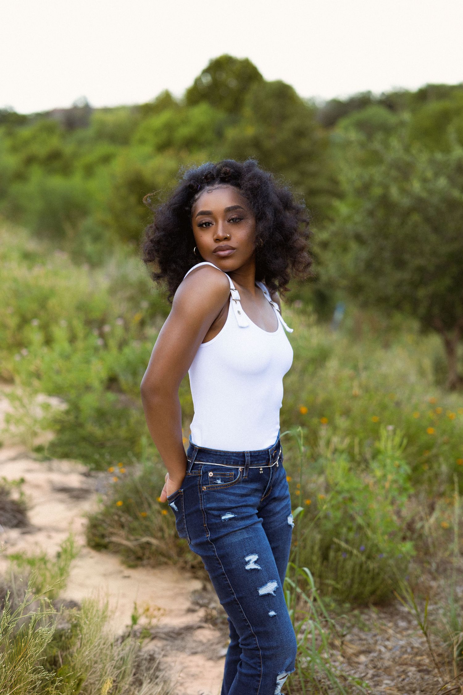 Young Black woman in white tank top and ripped jeans posing on a nature trail.