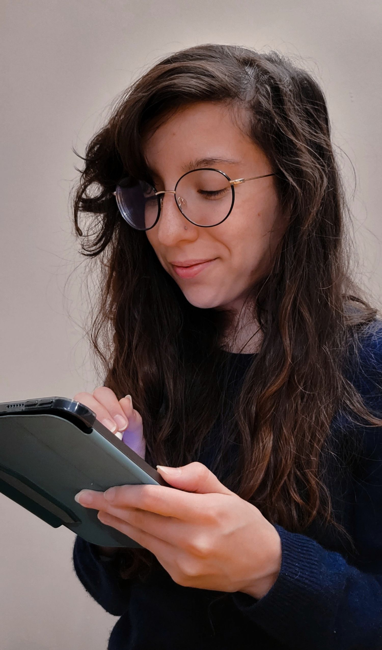 A young woman with long, dark hair and round glasses smiles while looking down at the tablet she is holding.