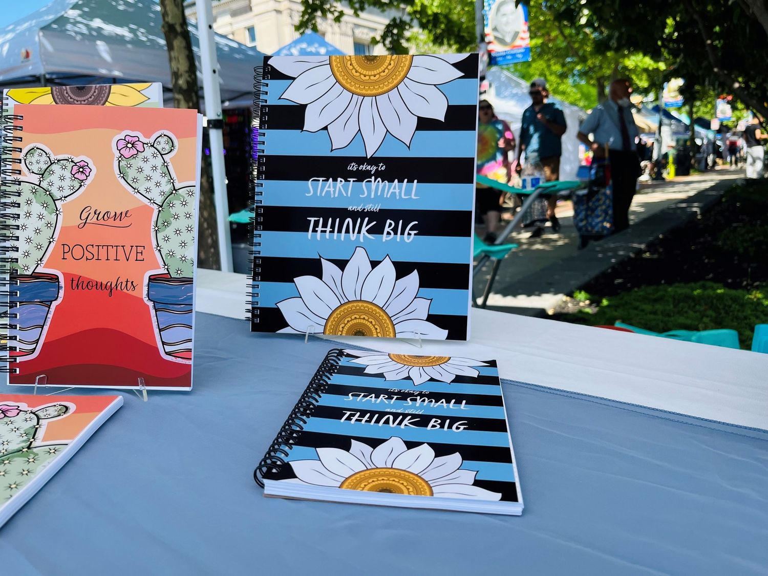 Spiral-bound notebooks with daisy and cactus designs on display at a market.