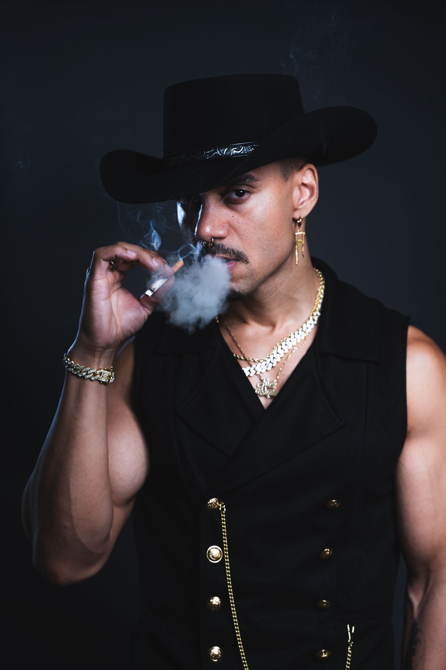 A stylish figure in a black cowboy hat and vest with gold jewelry exhales smoke while posing against a dark background.