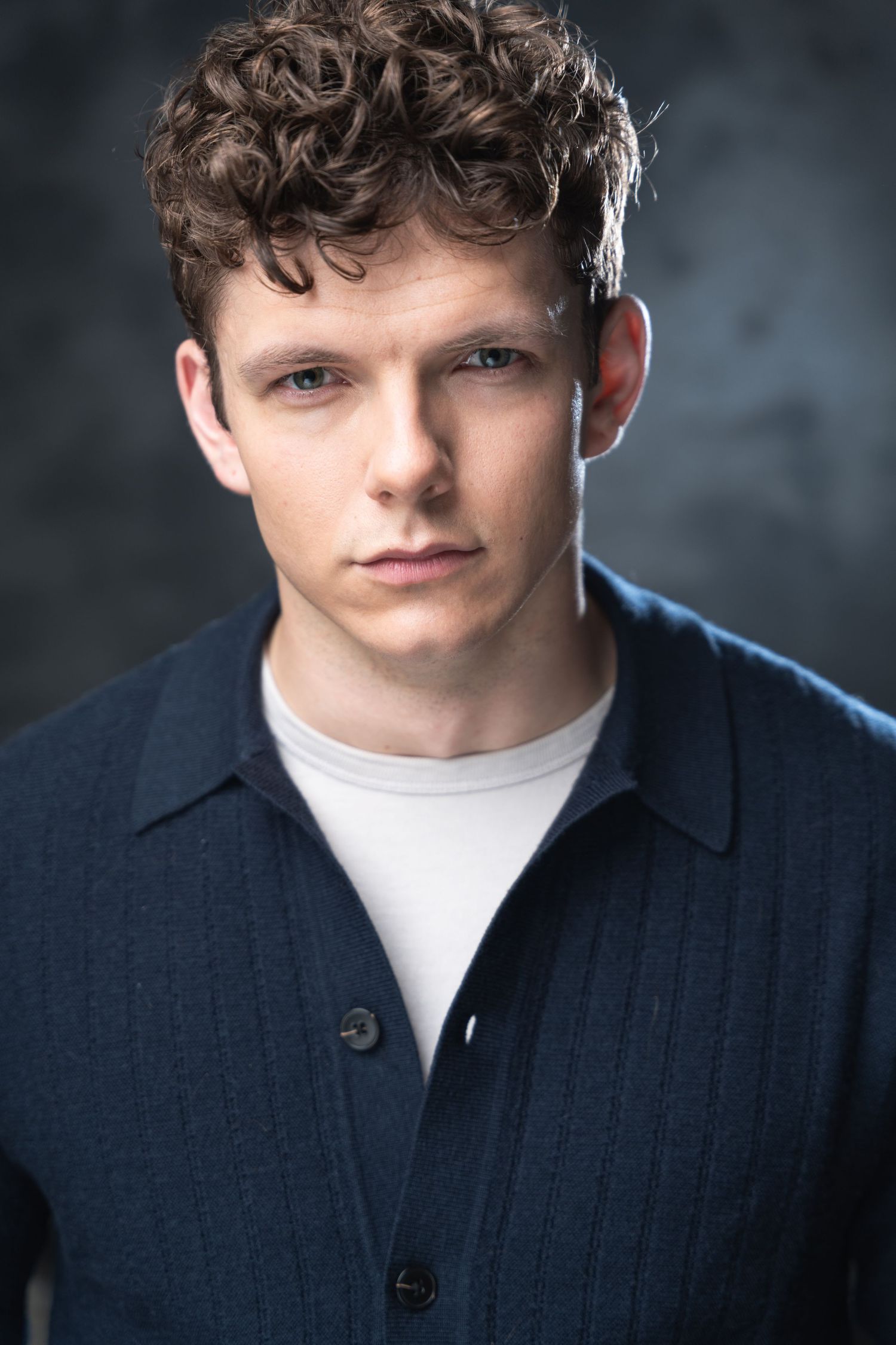 Portrait shot of a young person with curly hair wearing a navy button-up shirt over a white t-shirt against dark backdrop.