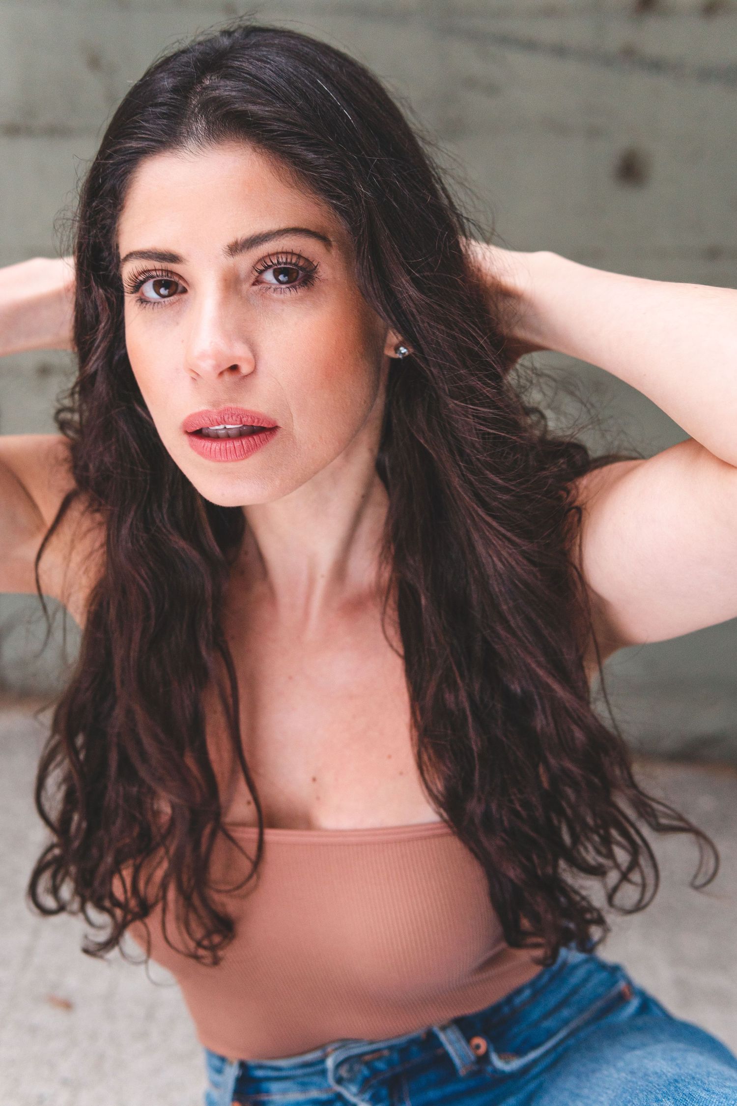 Portrait shot of someone in a nude colored top and denim, with long dark wavy hair against a textured backdrop.