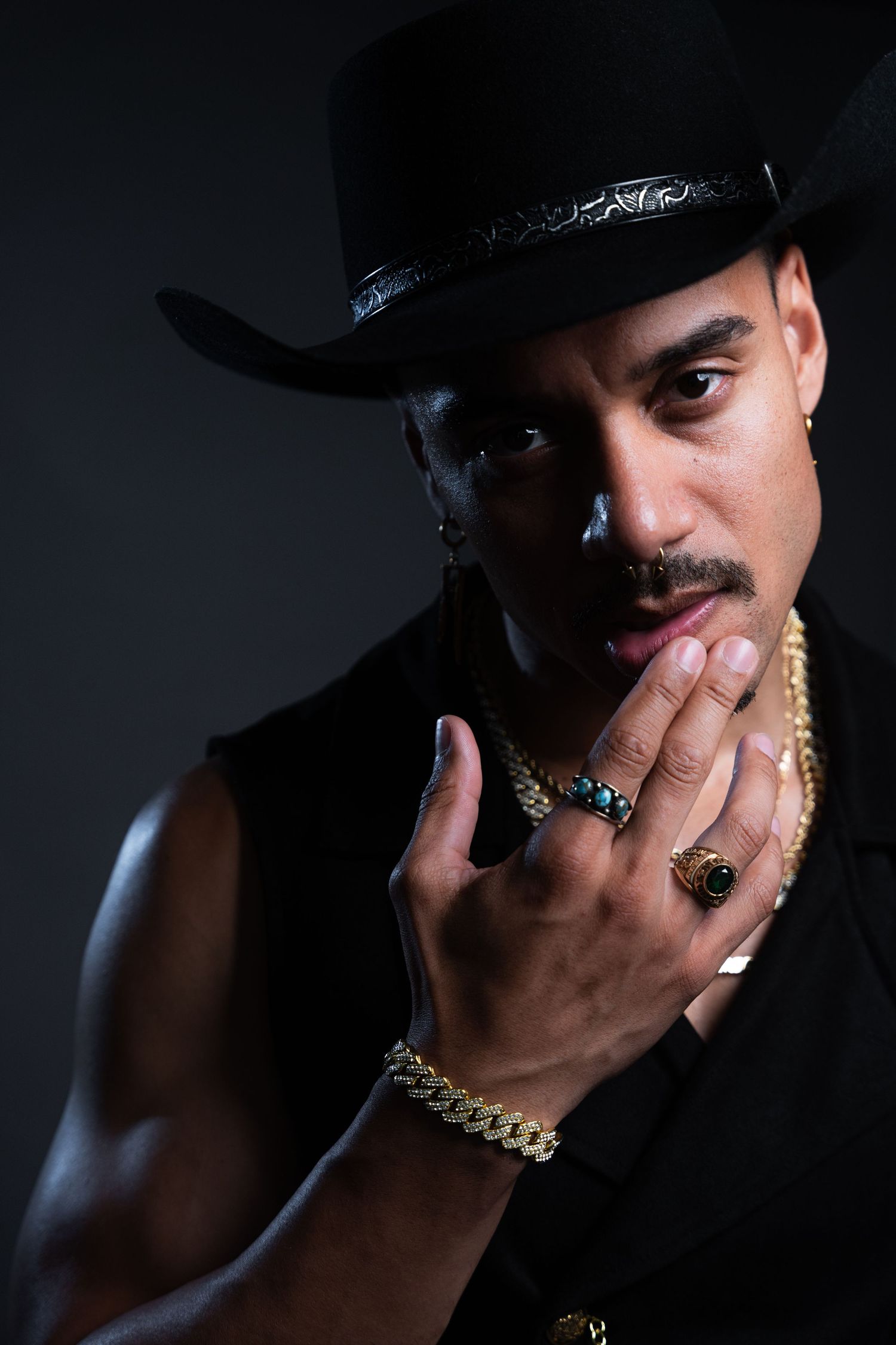 Portrait of person in black cowboy hat with gold jewelry and rings against dark background.