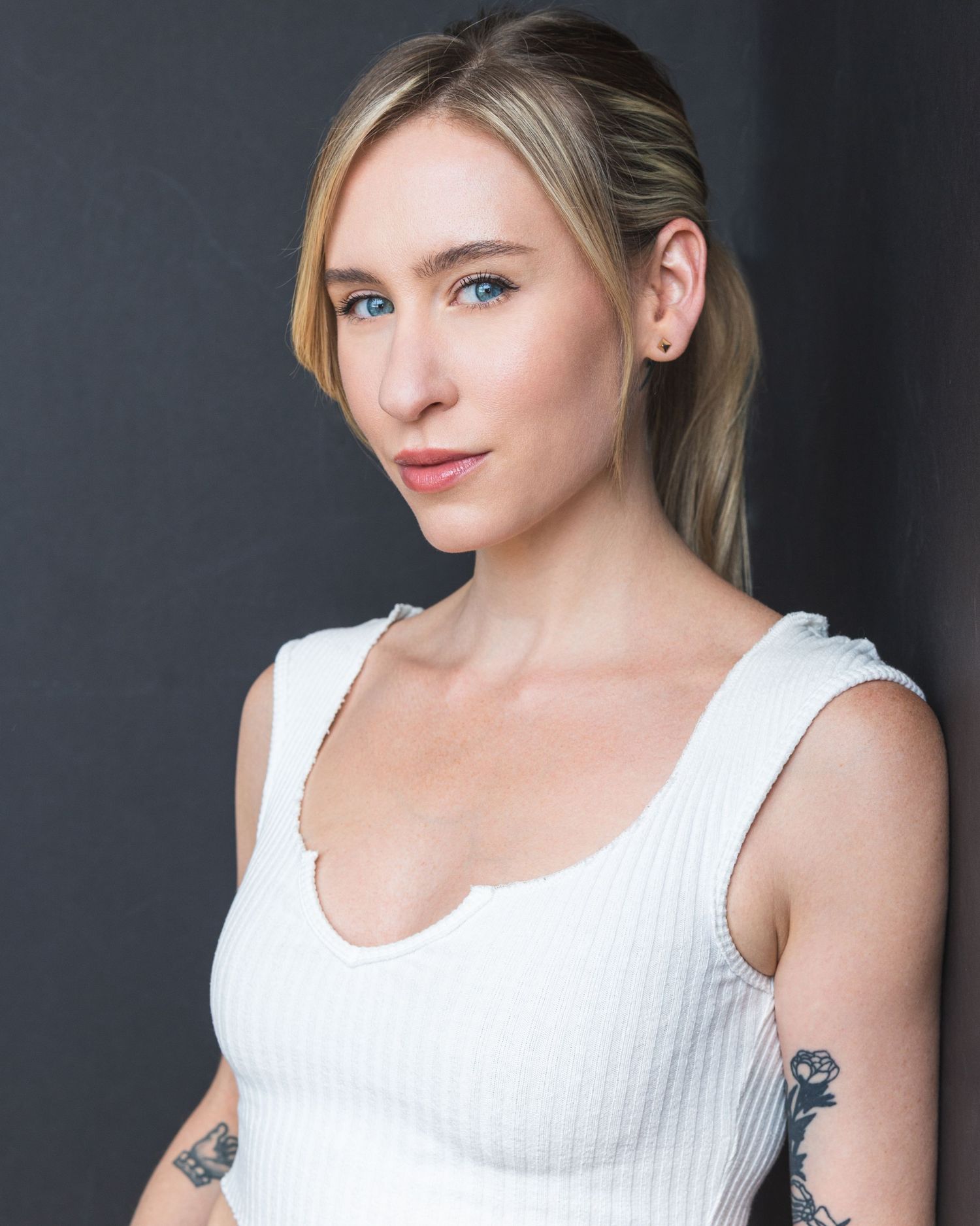 Portrait against dark background featuring someone with blonde hair in a white tank top showing visible tattoo on arm.