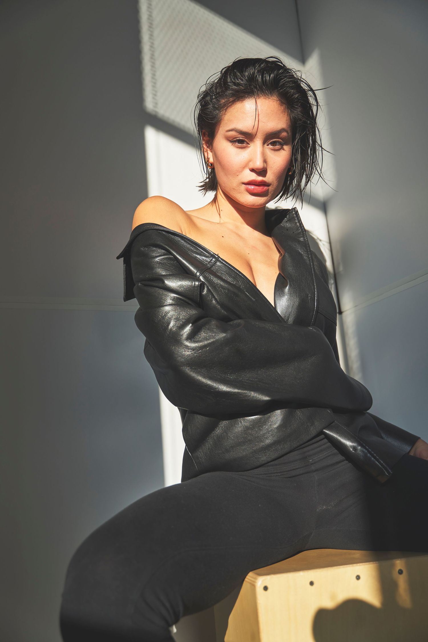 Fashion portrait with dramatic lighting shows model in black leather jacket against grey wall and natural window light.